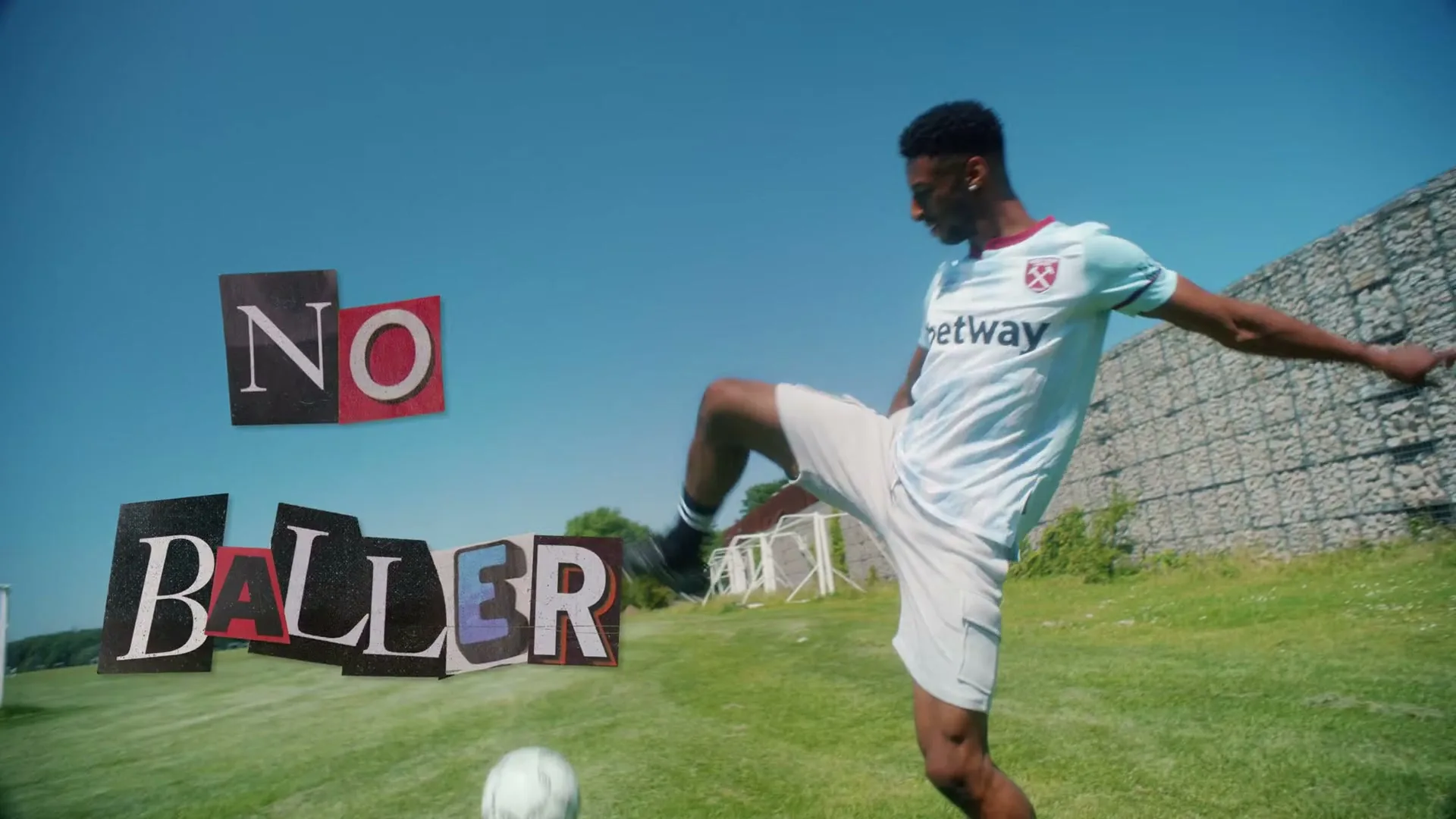 A young Black man in a white football kit juggles a football on a sunny day, with "NO BALLER" text floating beside him.