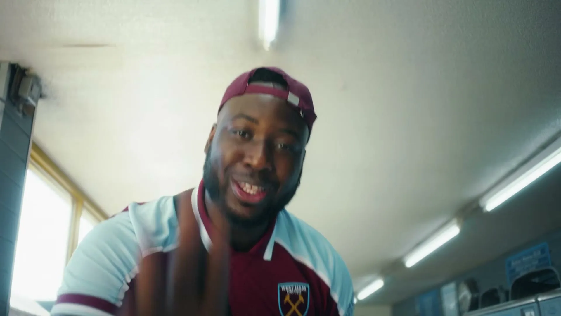 A Black man in his 20s, wearing a West Ham United football jersey and a cap, gestures with his hands in a laundromat.