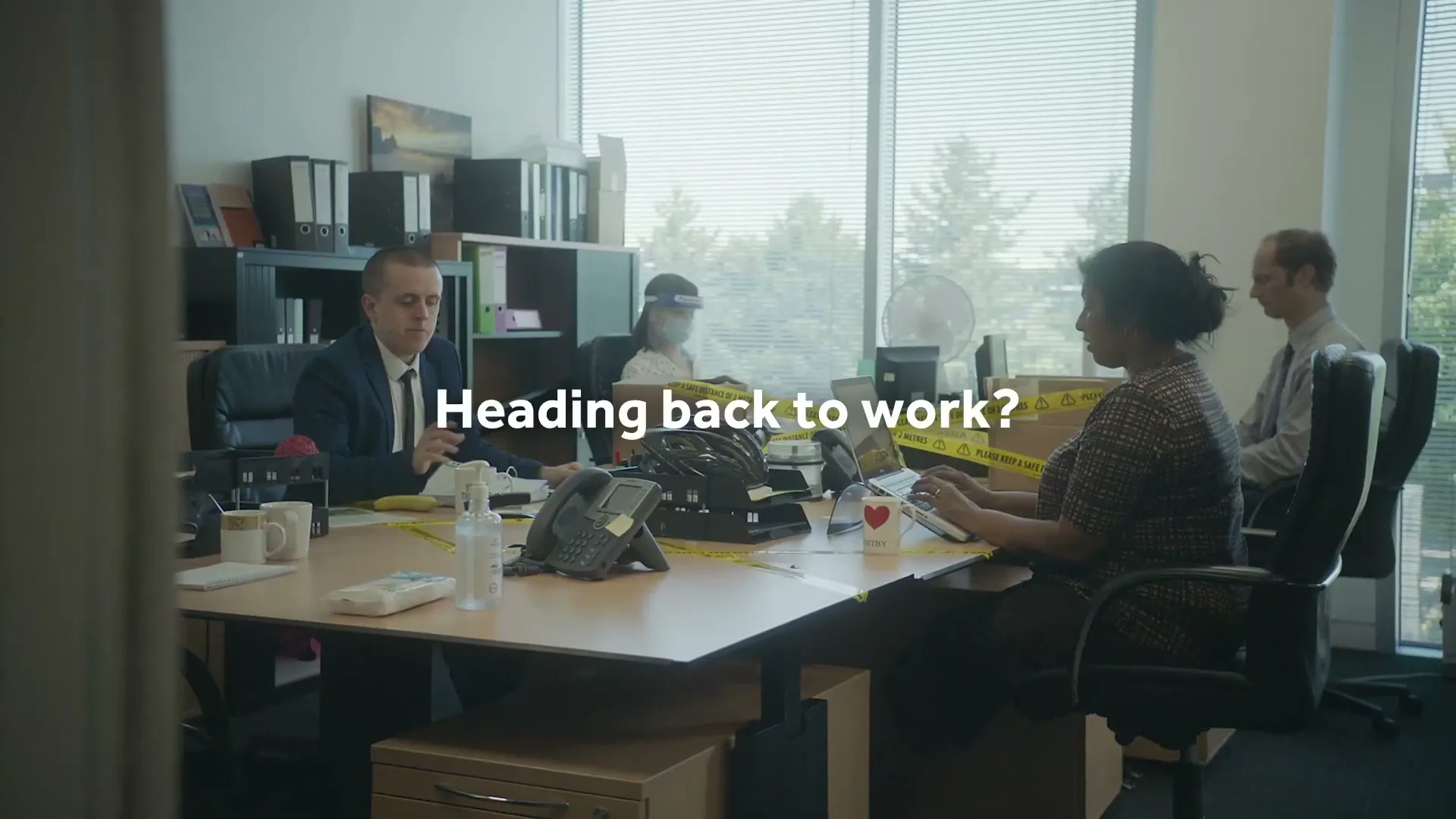 Four office workers are seen in an open-plan office, with yellow caution tape marking social distancing around their desks.