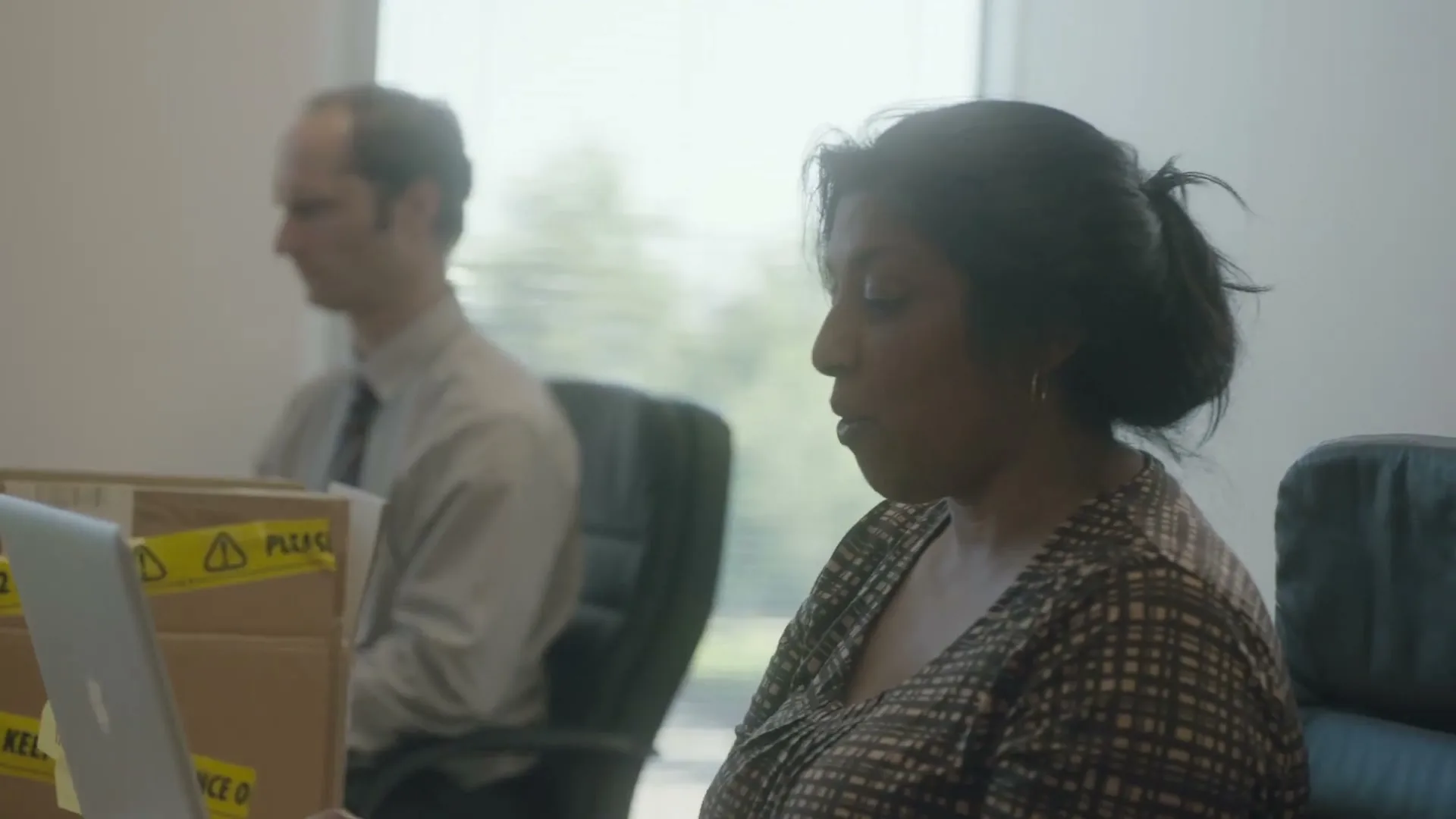 A woman with dark hair in a patterned top sits at a desk, looking down at a laptop, with a man in a shirt and tie blurred in the background.