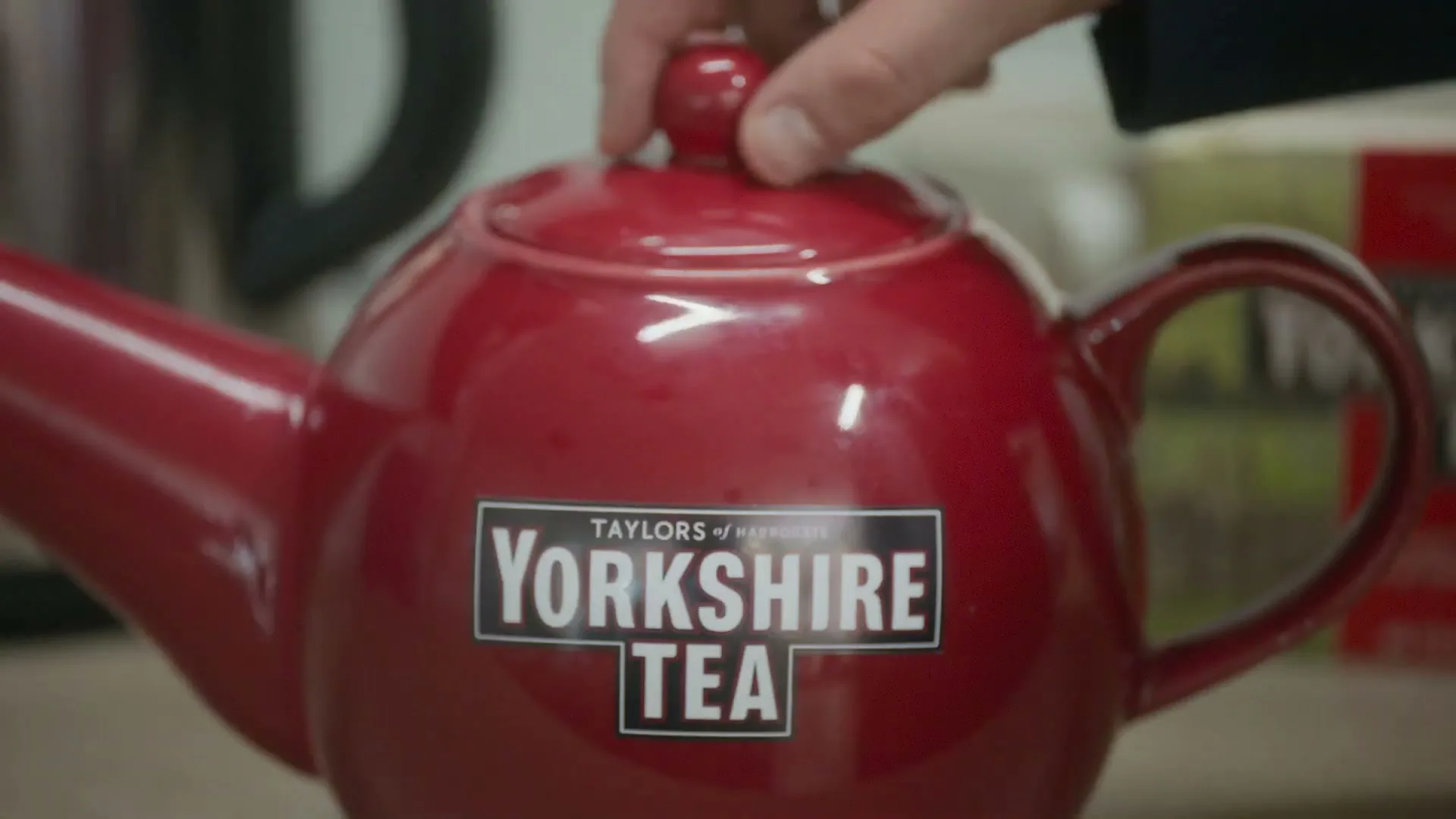 A hand removes the lid from a red Yorkshire Tea teapot, revealing the opening, in a kitchen setting.