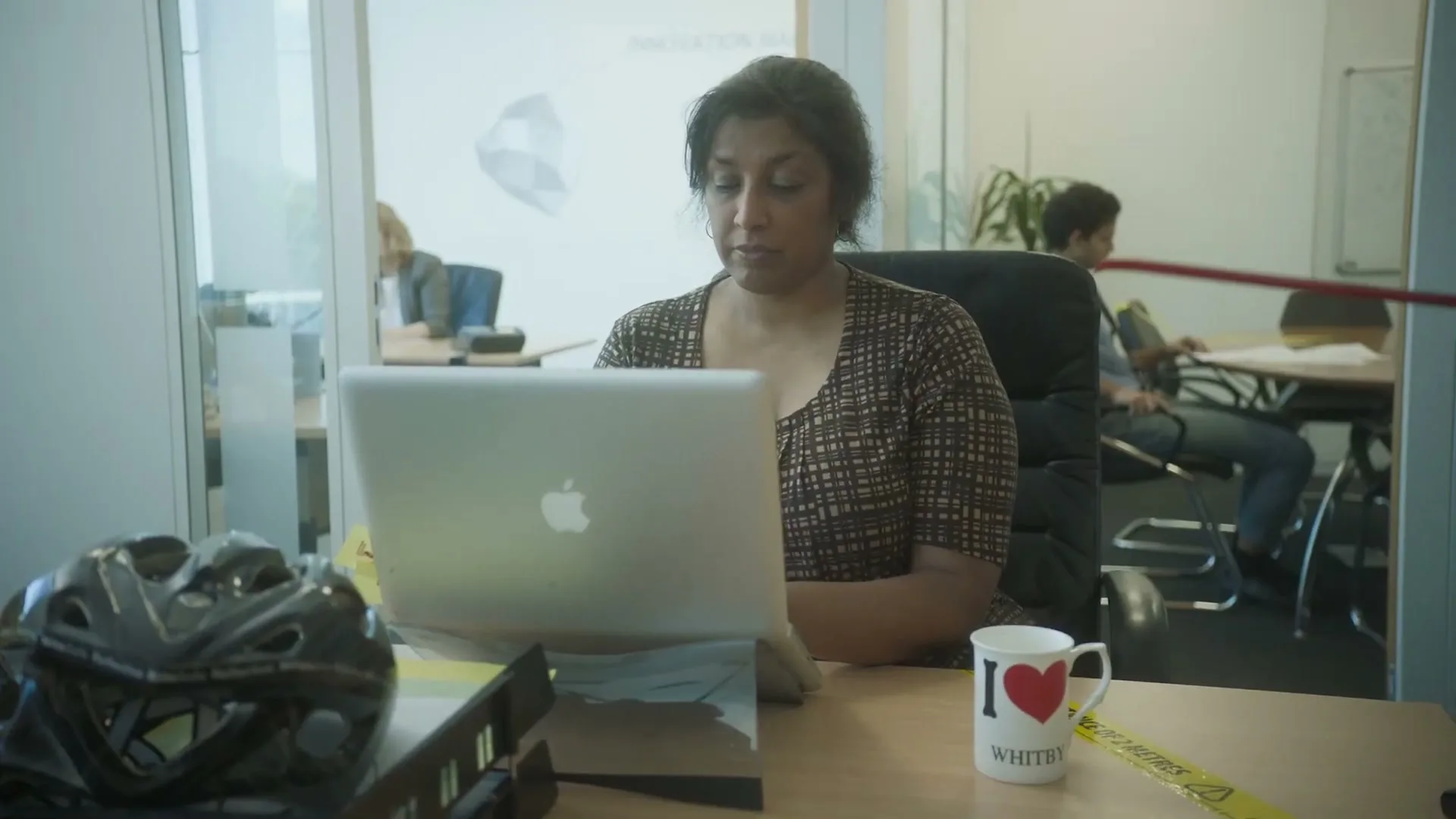 A woman with dark hair in a patterned top works on a laptop at a desk in an office, while a red pole extends across the frame.