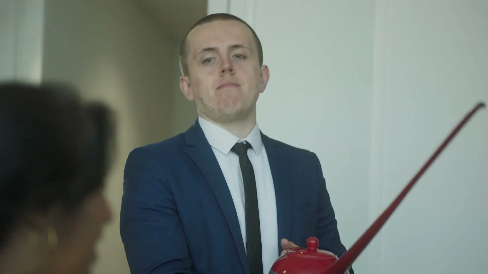 A young man in a blue suit holds a red teapot with an extremely long spout, looking at a woman off-screen in an office.