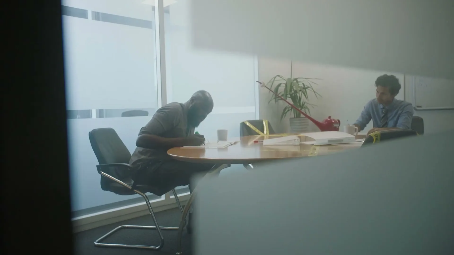 Two men are seen through a glass partition in an office, one writing at a table and the other looking at papers.