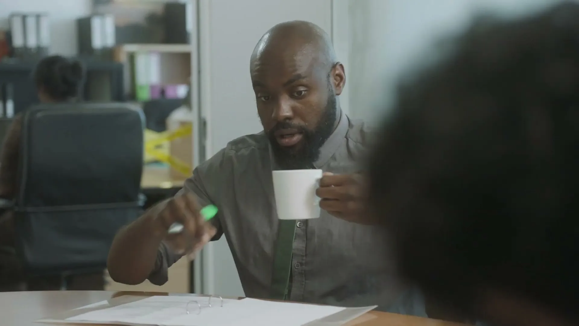 A bald Black man with a beard, wearing a grey shirt and green tie, holds a white mug and a green pen while looking at papers on a desk in an office.