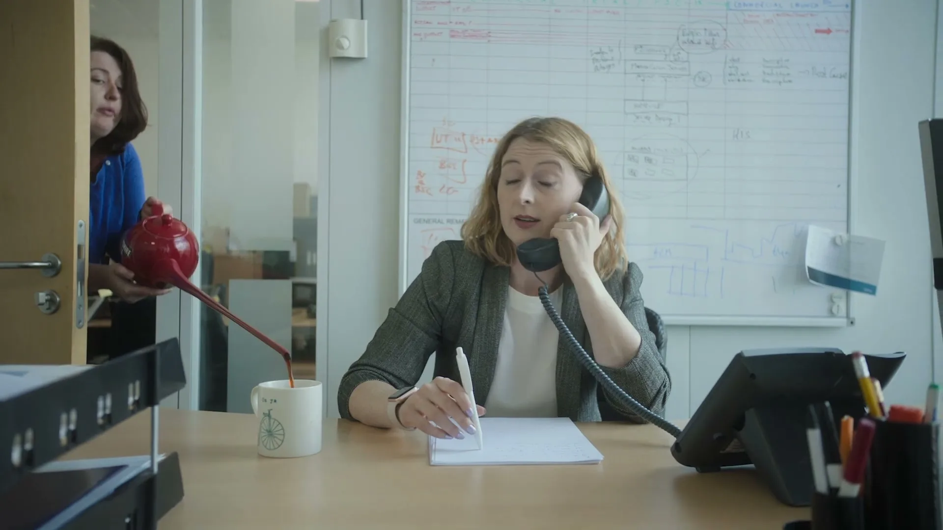 A woman in a grey blazer talks on a landline phone at her office desk, while another woman pours tea from a red teapot into a mug from behind a door.