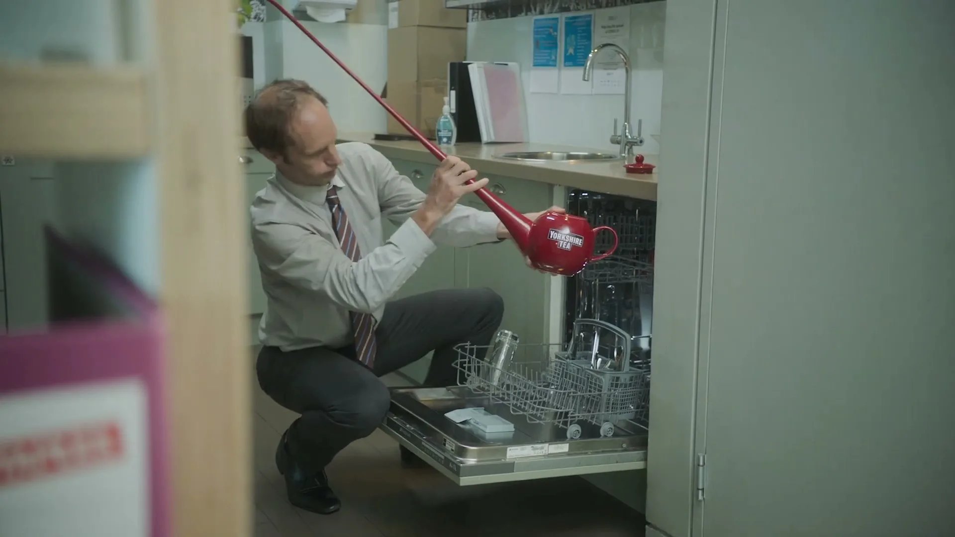 A man in a shirt and tie uses a long red pole to place a red teapot into a dishwasher in an office kitchen.