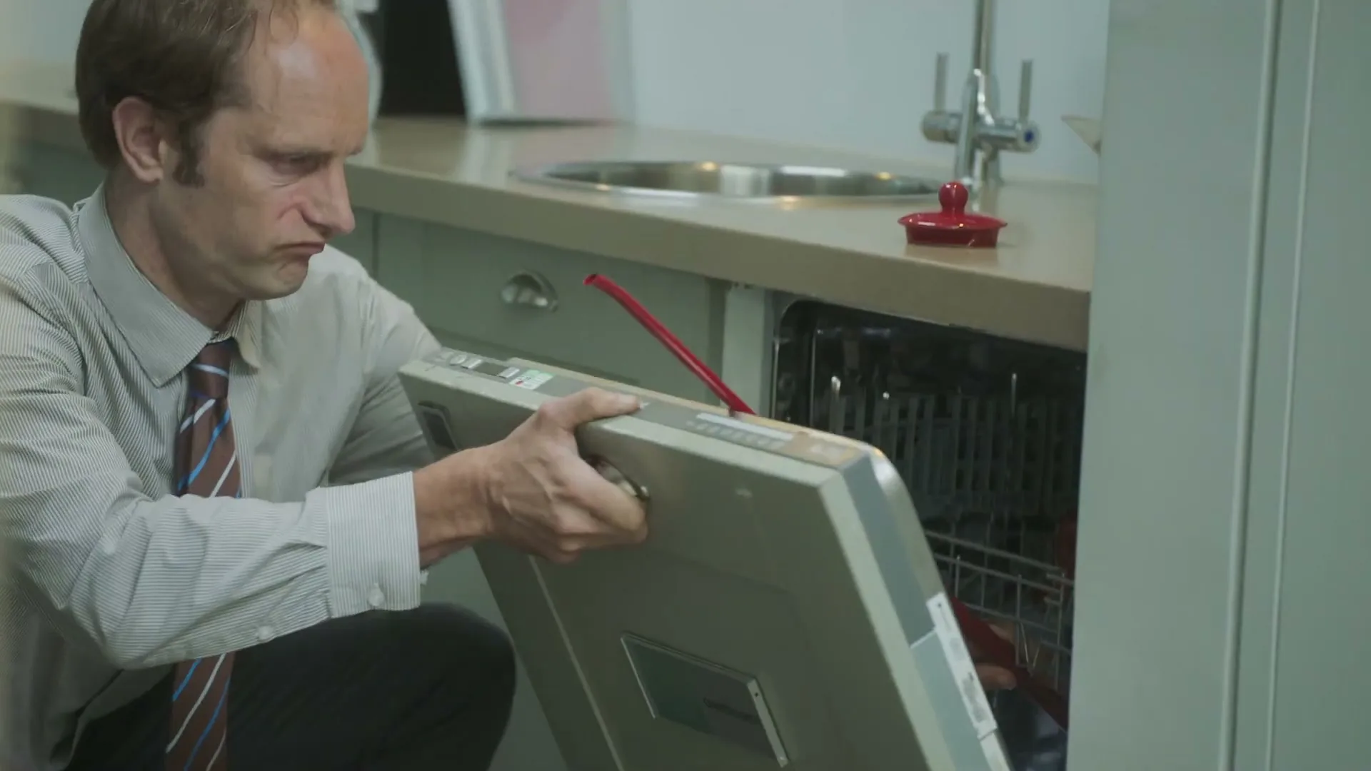 A man in a shirt and tie struggles to fit a large, flat object into a dishwasher in a modern kitchen.