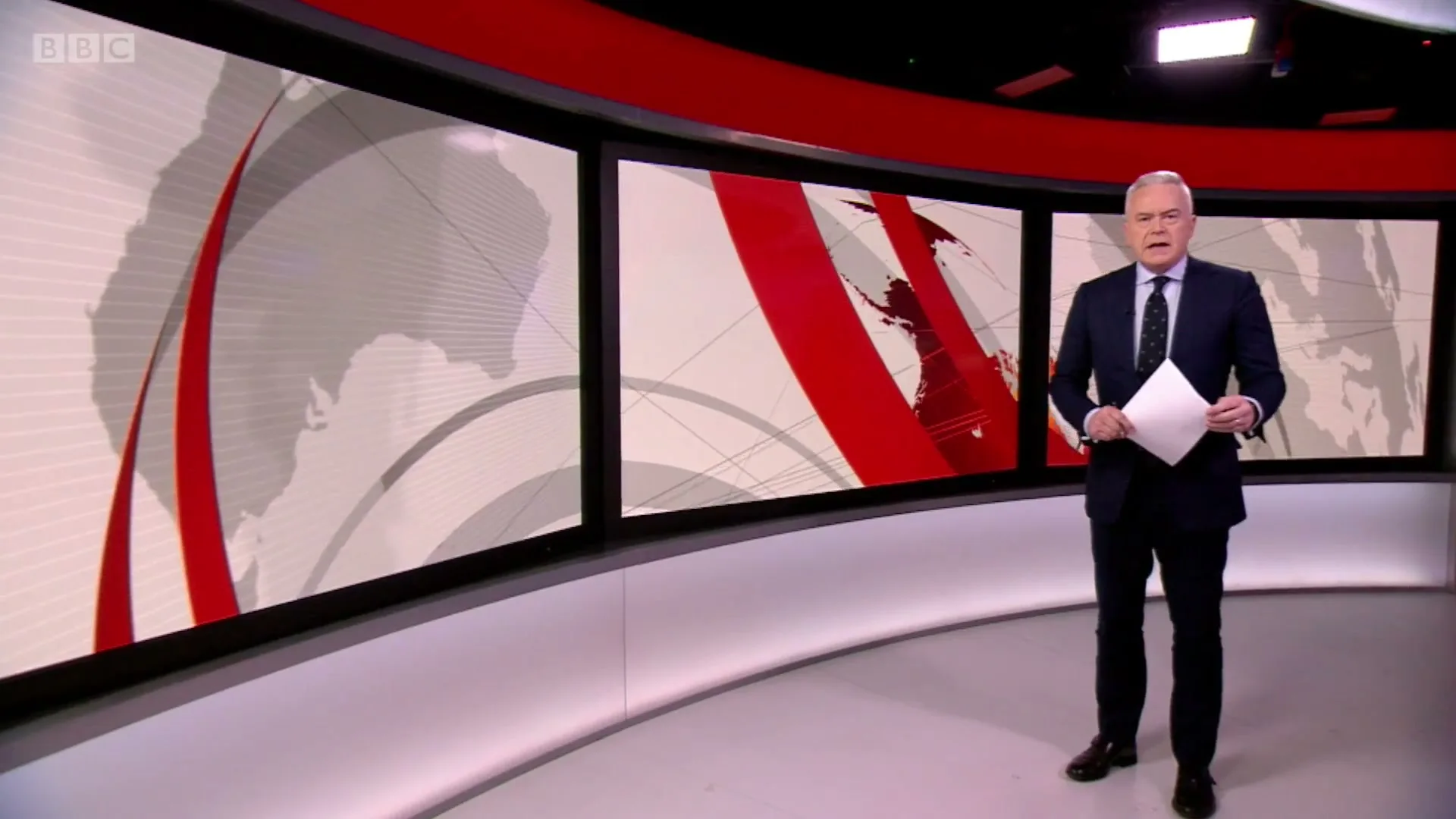A male news anchor in a suit stands in a brightly lit news studio with large screens displaying a world map and red graphics.