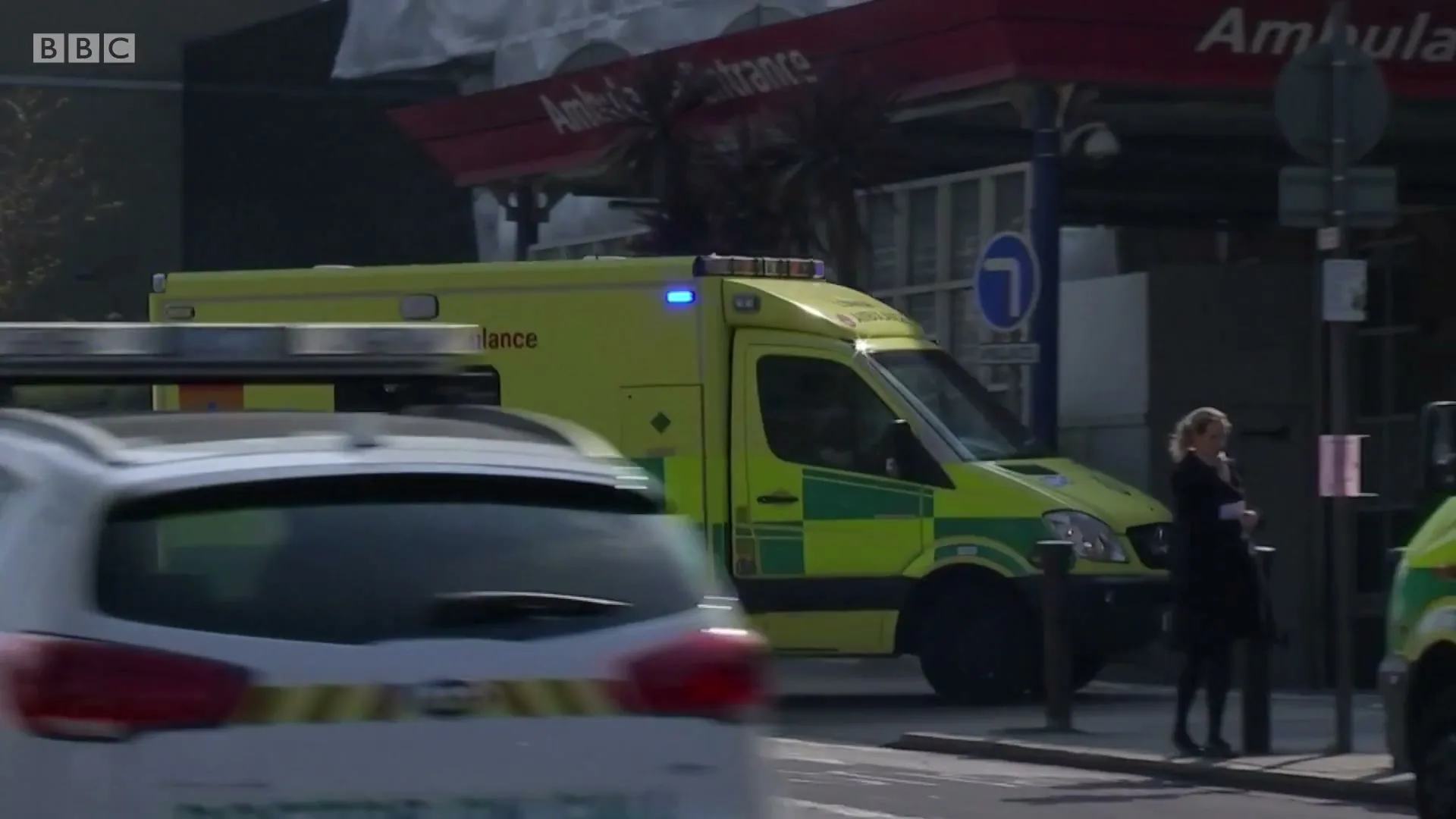 An ambulance with flashing blue lights is parked outside a hospital entrance, while a woman stands nearby and a white car drives past.