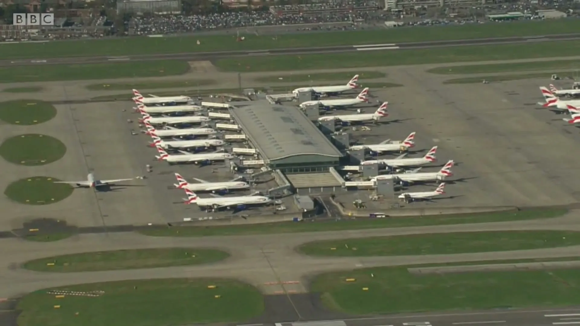 An aerial view shows numerous British Airways planes parked at an airport terminal on a sunny day, with a distant city visible.