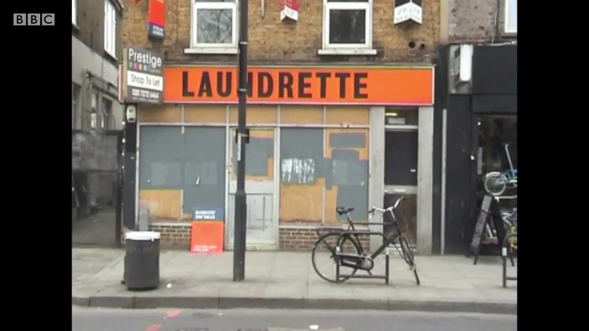 A wide shot of a boarded-up laundrette with an orange sign, a bicycle, and a trash can on the sidewalk in an urban setting.