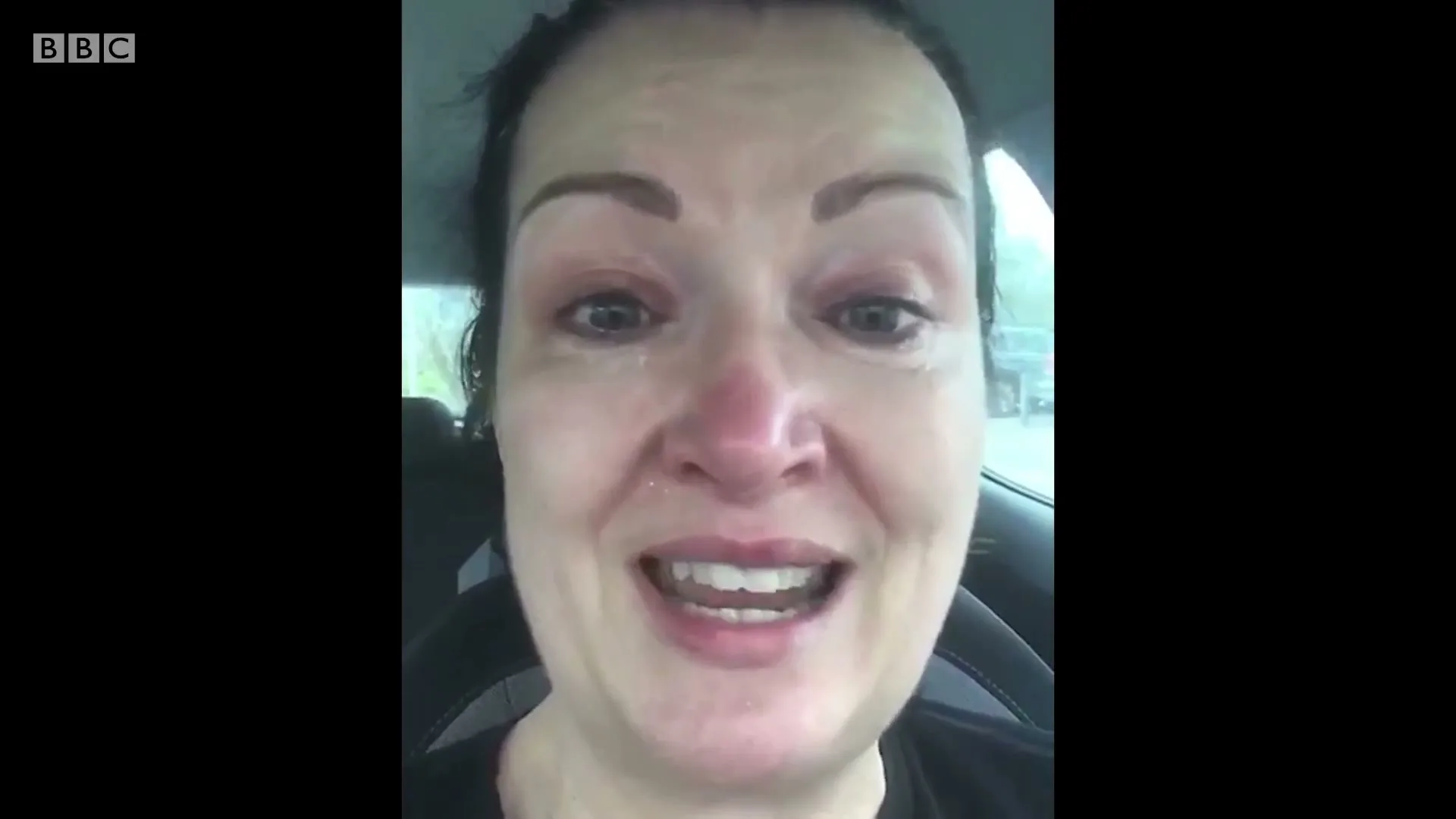 A woman with dark hair and a black shirt cries while speaking to the camera from inside a car.