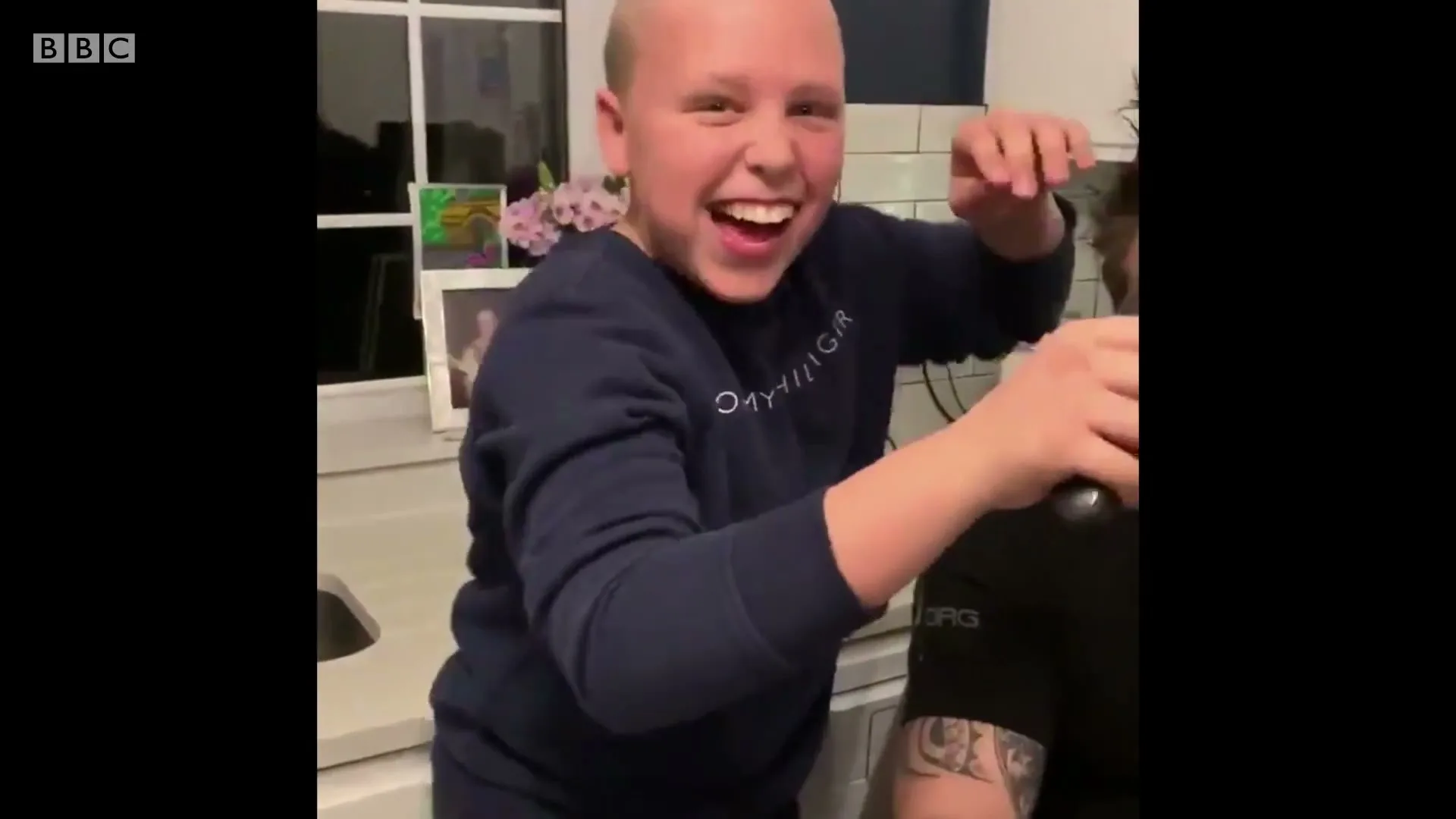A bald child in a blue sweatshirt laughs while shaving the side of an adult's head in a kitchen.