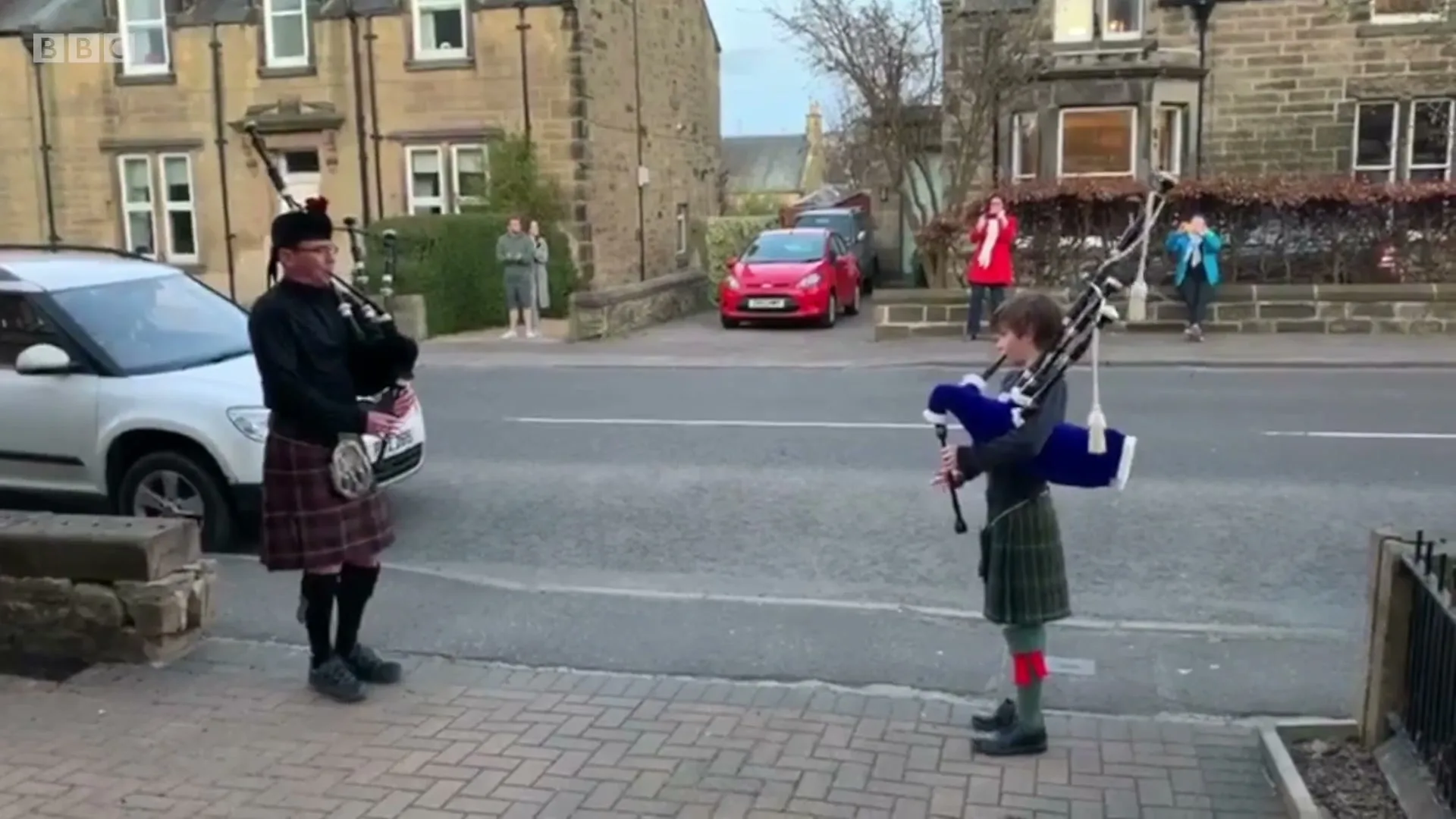 A man and a child in kilts play bagpipes across a street from each other, while neighbors watch from their homes.