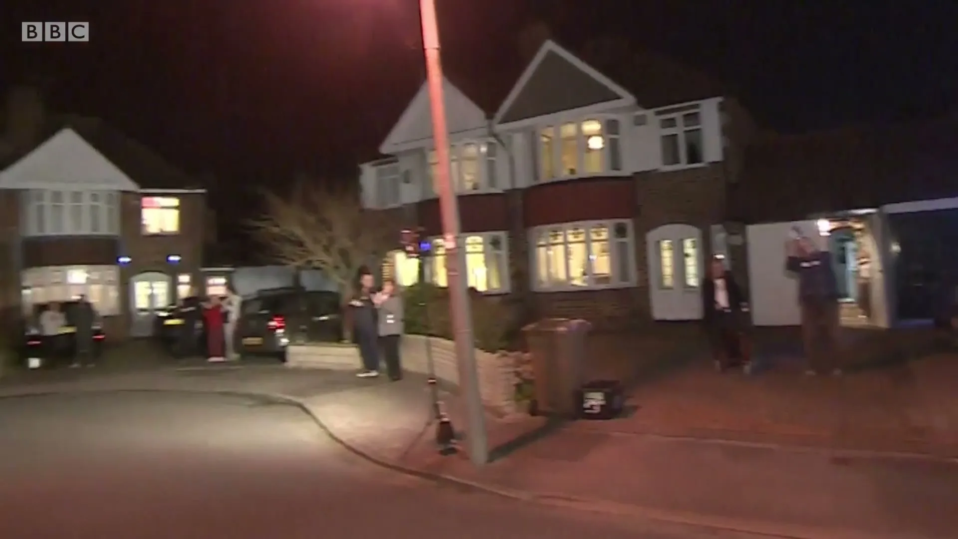 Neighbors stand outside their homes at night, clapping and cheering in a suburban street, illuminated by streetlights and house windows.