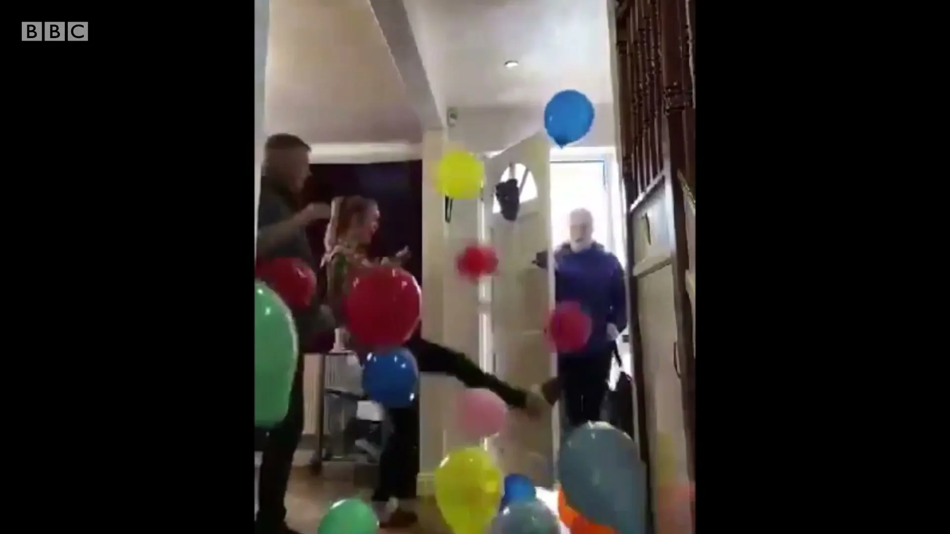A family surprises an older woman with balloons as she enters a home, creating a joyful and heartwarming moment.