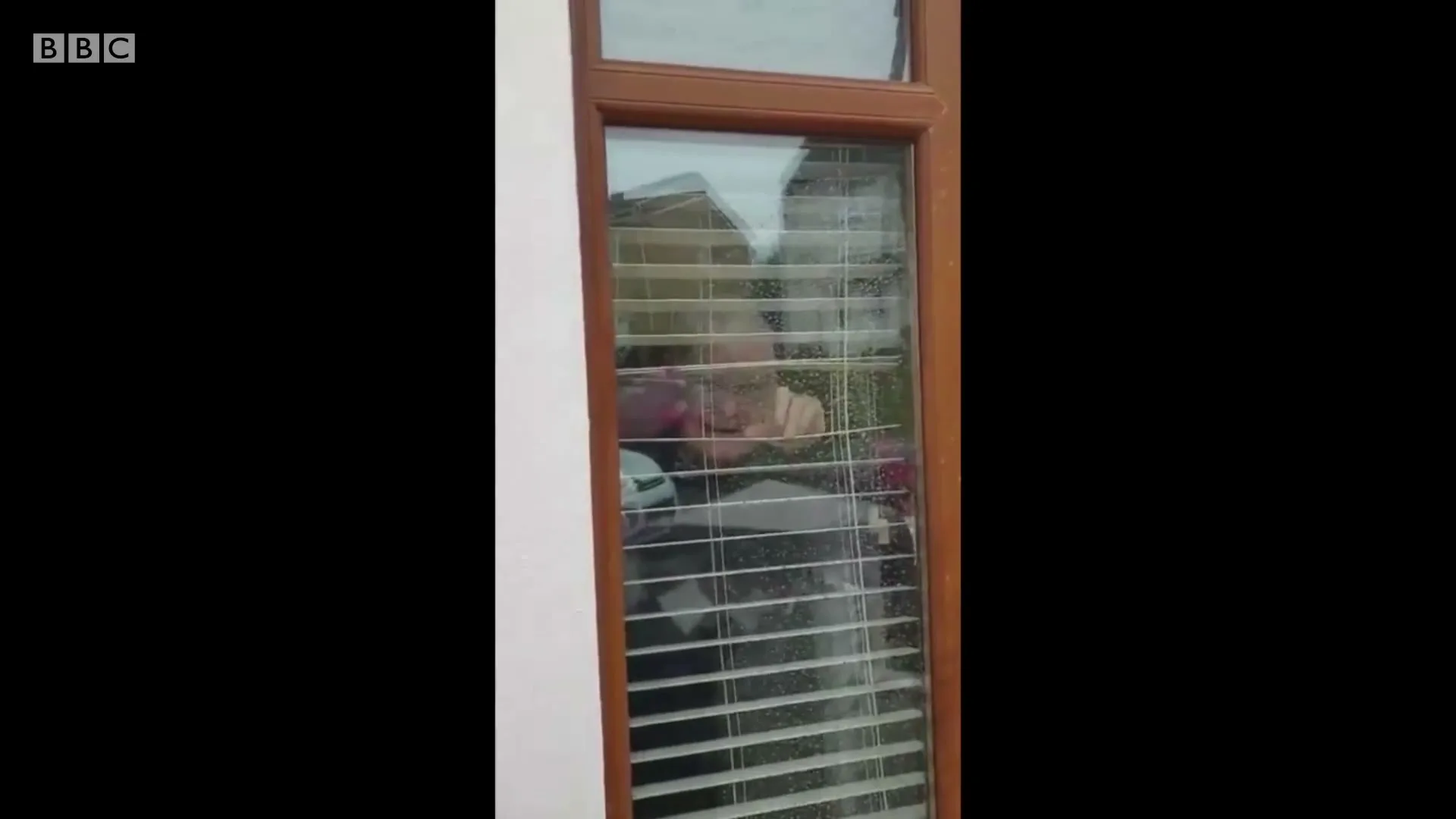 A child with blonde hair looks out a window with blinds, their face partially obscured, appearing to be inside a home during the day.