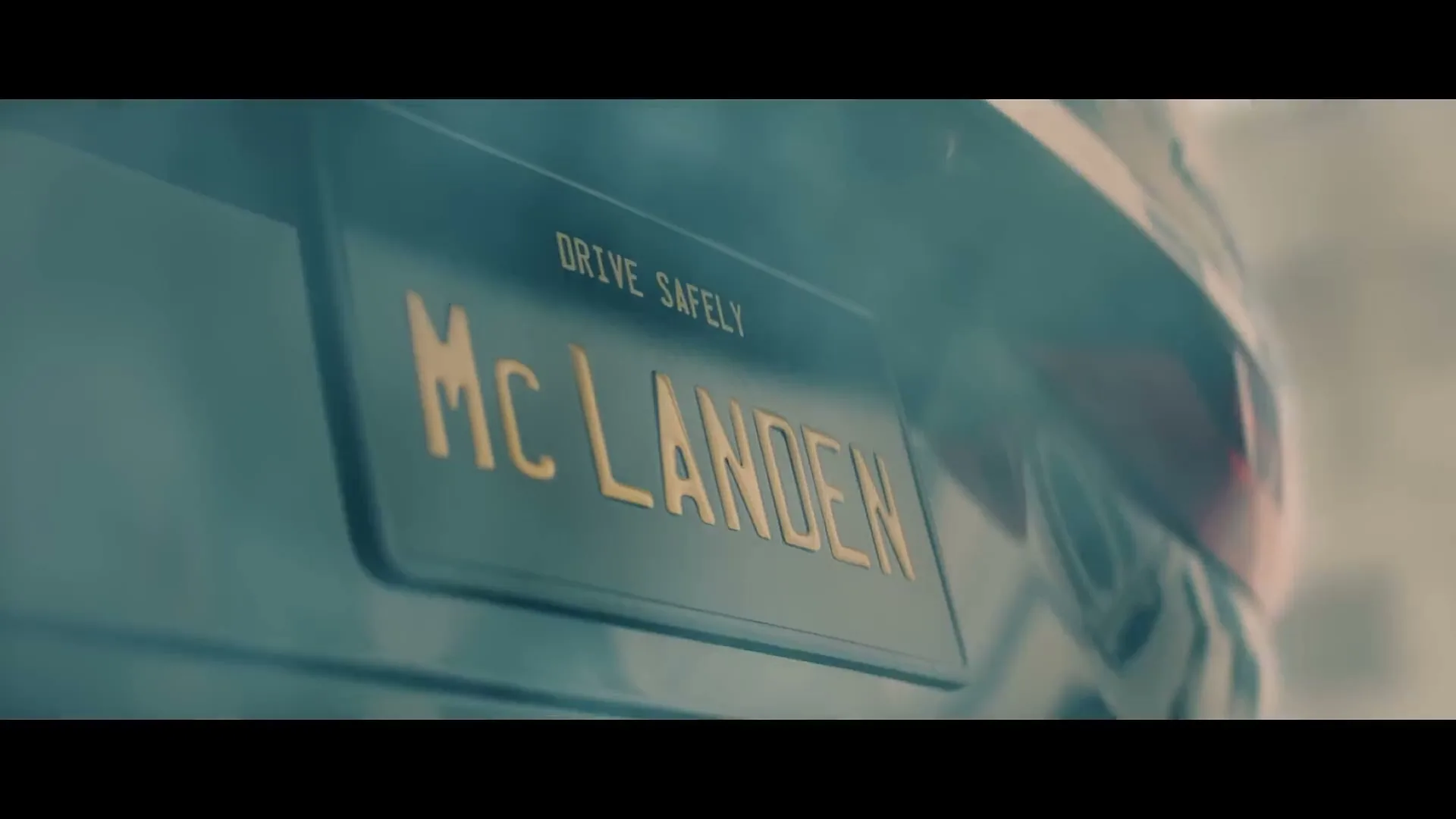 A close-up shot of a car's license plate, which reads "McLANDEN" in gold letters and "DRIVE SAFELY" above it, is shown.