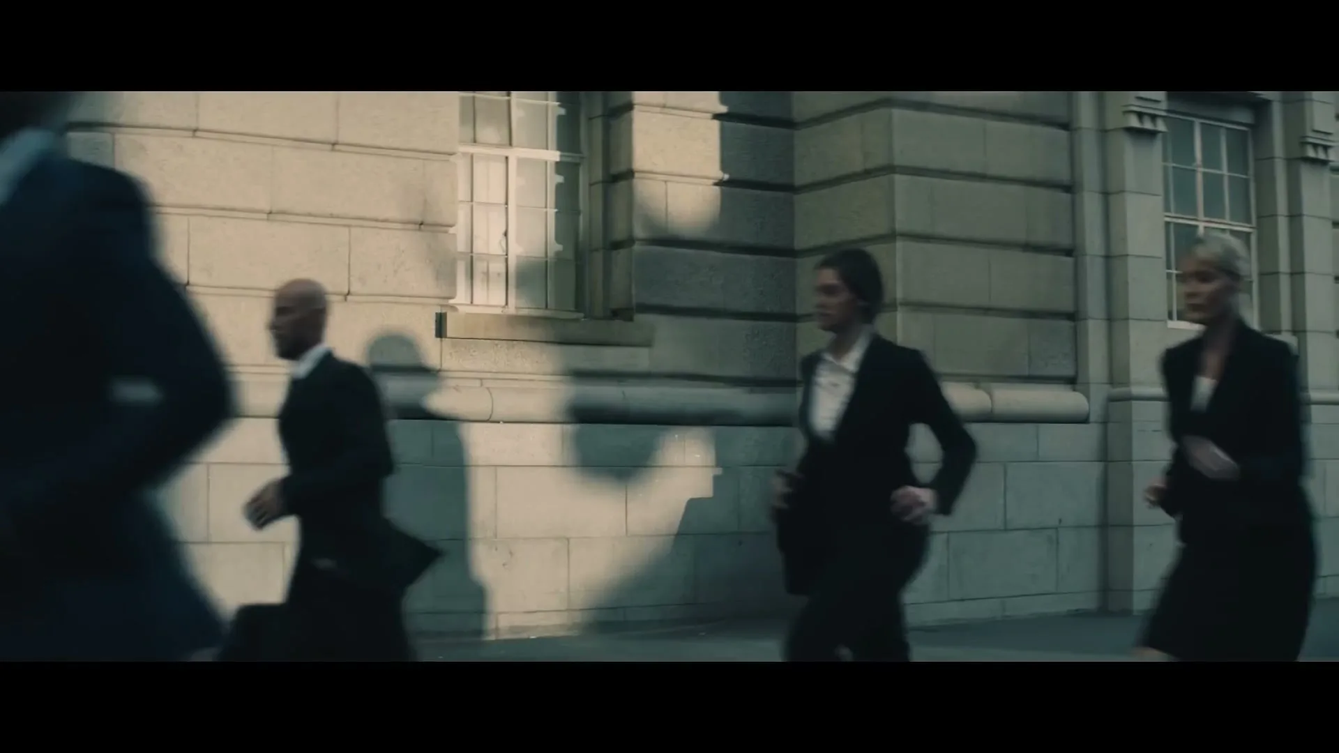 A diverse group of business people in suits run past a stone building with windows, casting long shadows on the wall.
