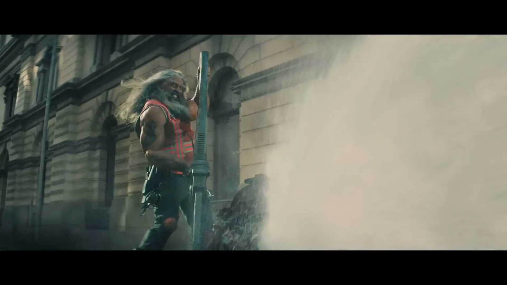 A muscular, long-haired man with a beard, wearing a plaid vest, stands on a city street, holding a large pipe as water sprays from a broken fire hydrant.