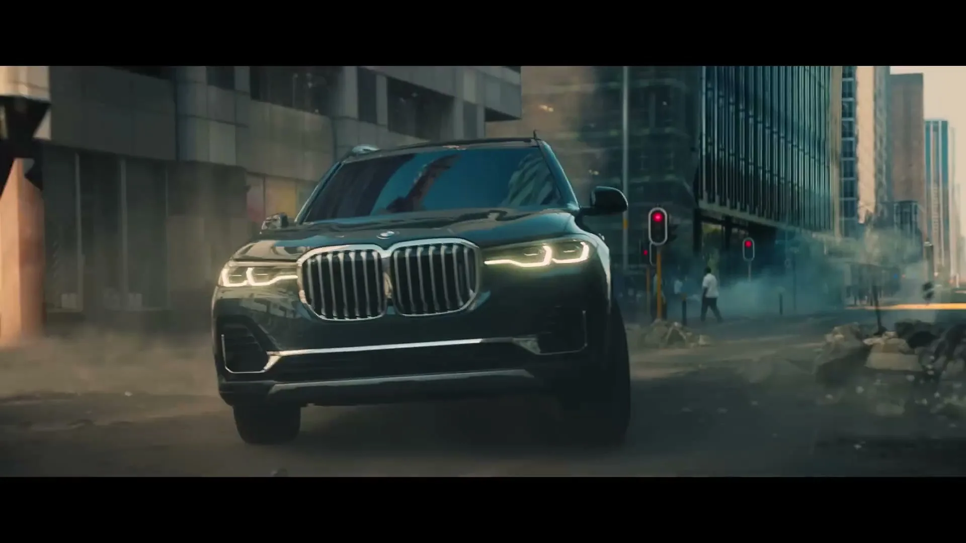 A dark BMW SUV drives through a dusty, debris-filled city street with tall buildings and a traffic light in the background.