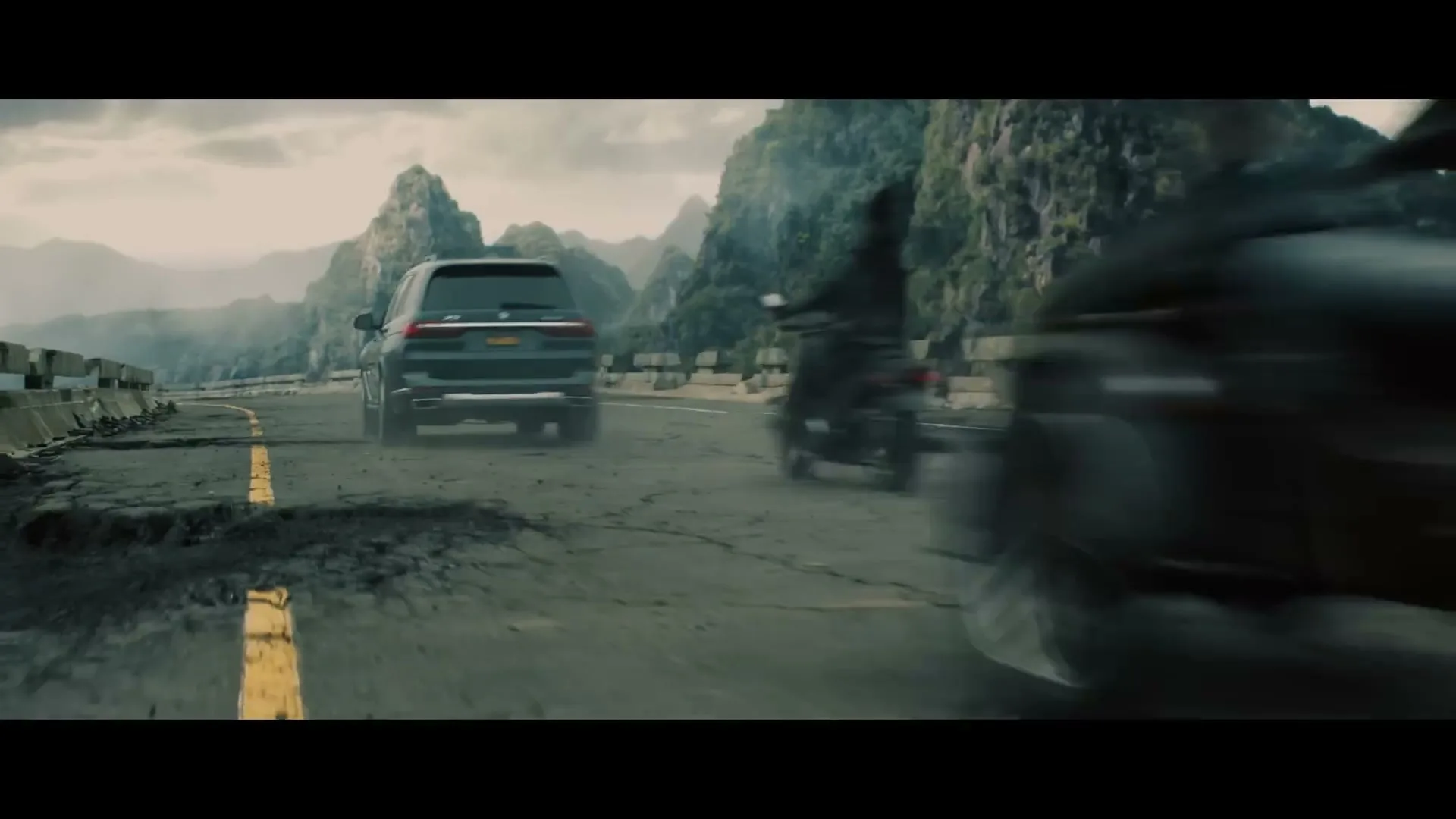 A dark SUV drives away on a cracked road, with a motorcycle passing on the right, against a backdrop of green mountains under a cloudy sky.
