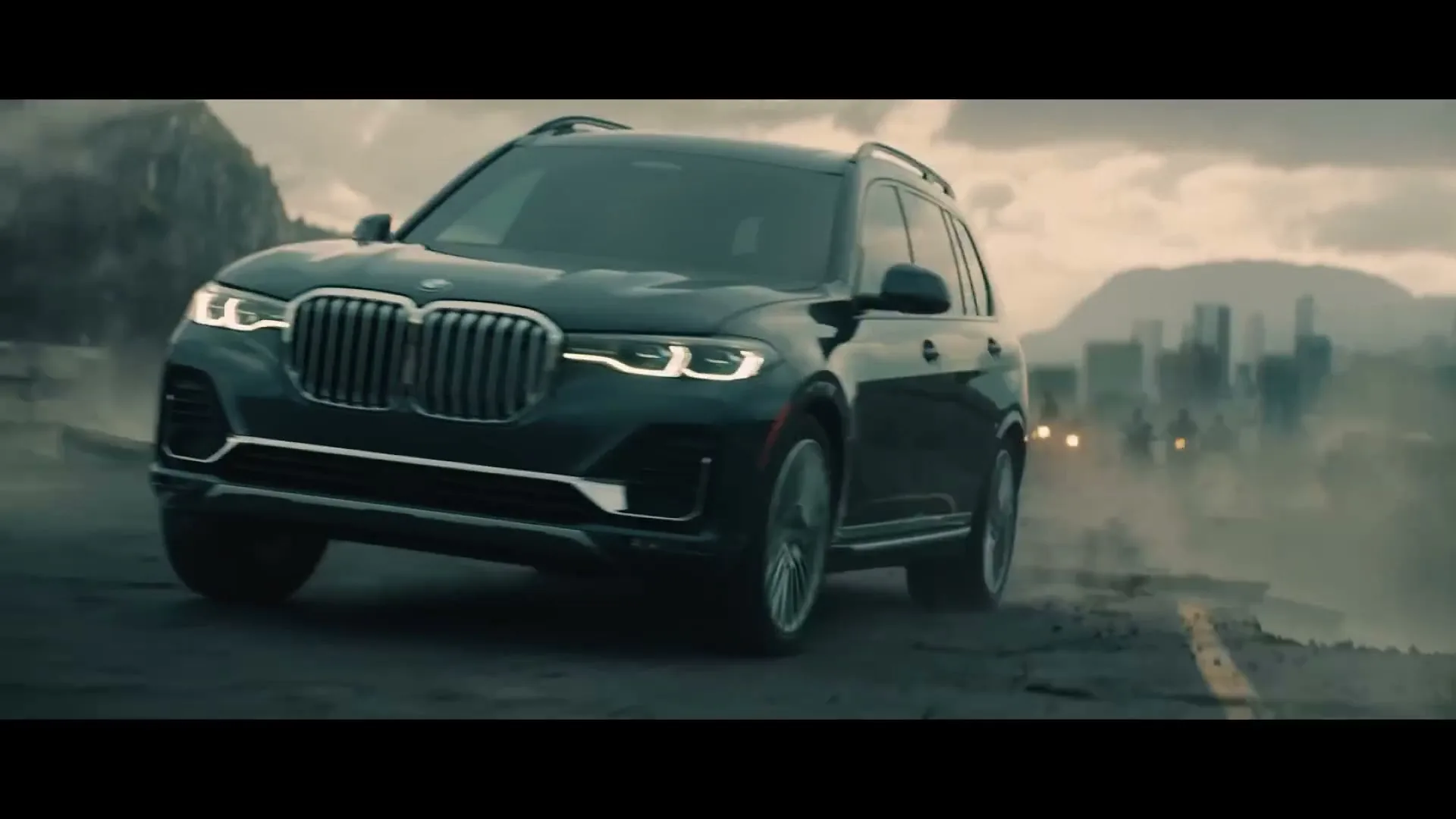 A dark BMW SUV drives on a cracked road, with a city skyline in the background under a cloudy sky.