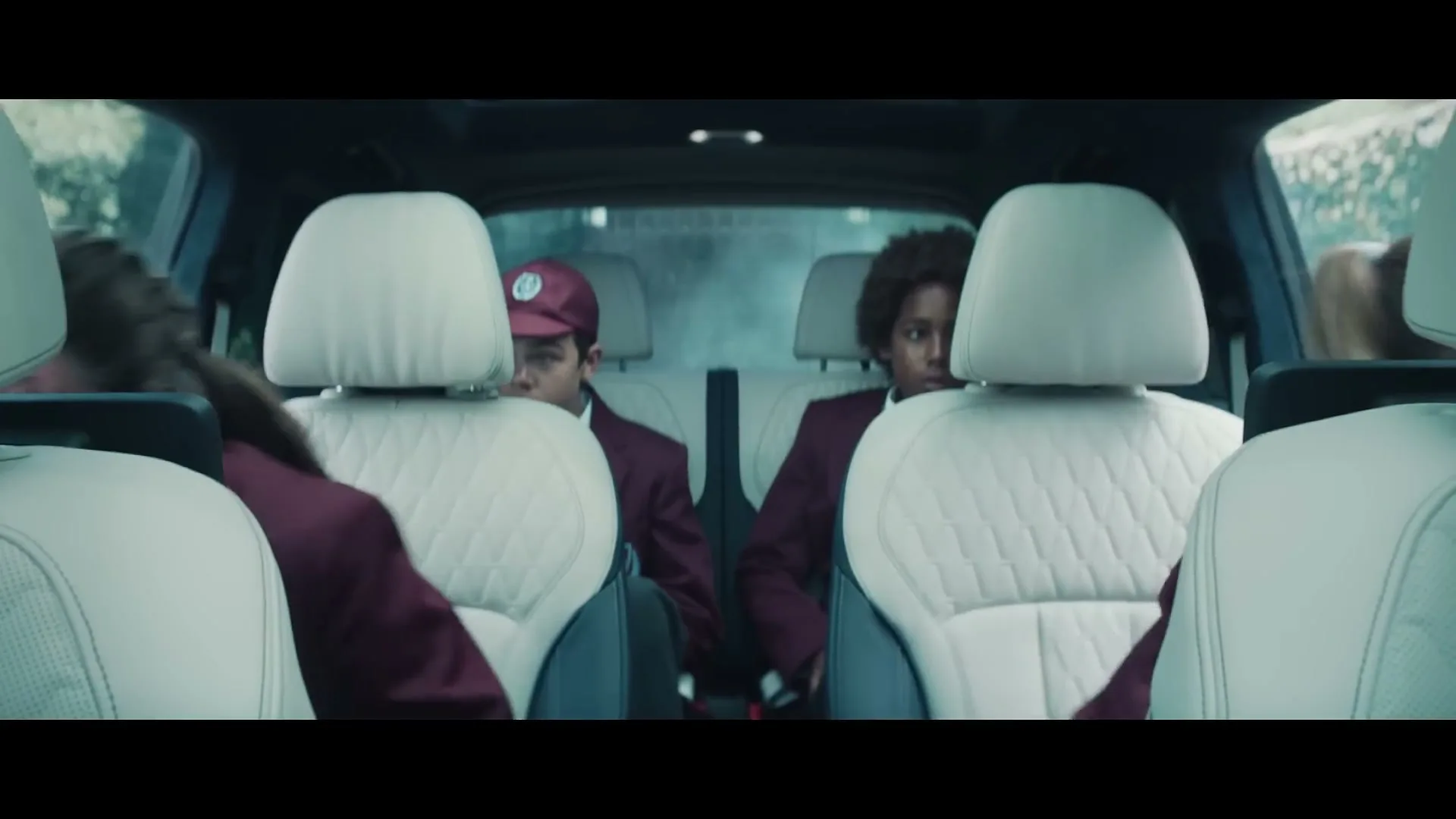 Four children in school uniforms are seen from behind the front seats of a car, looking around with surprised expressions as they are driven.