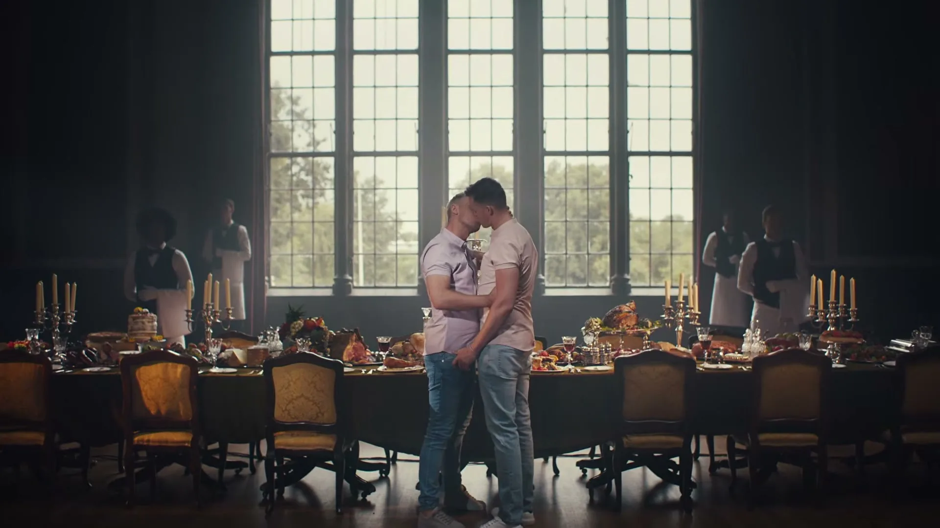 Two men in casual clothes kiss passionately at the head of a long, opulent dining table, surrounded by formally dressed servers in a grand hall.