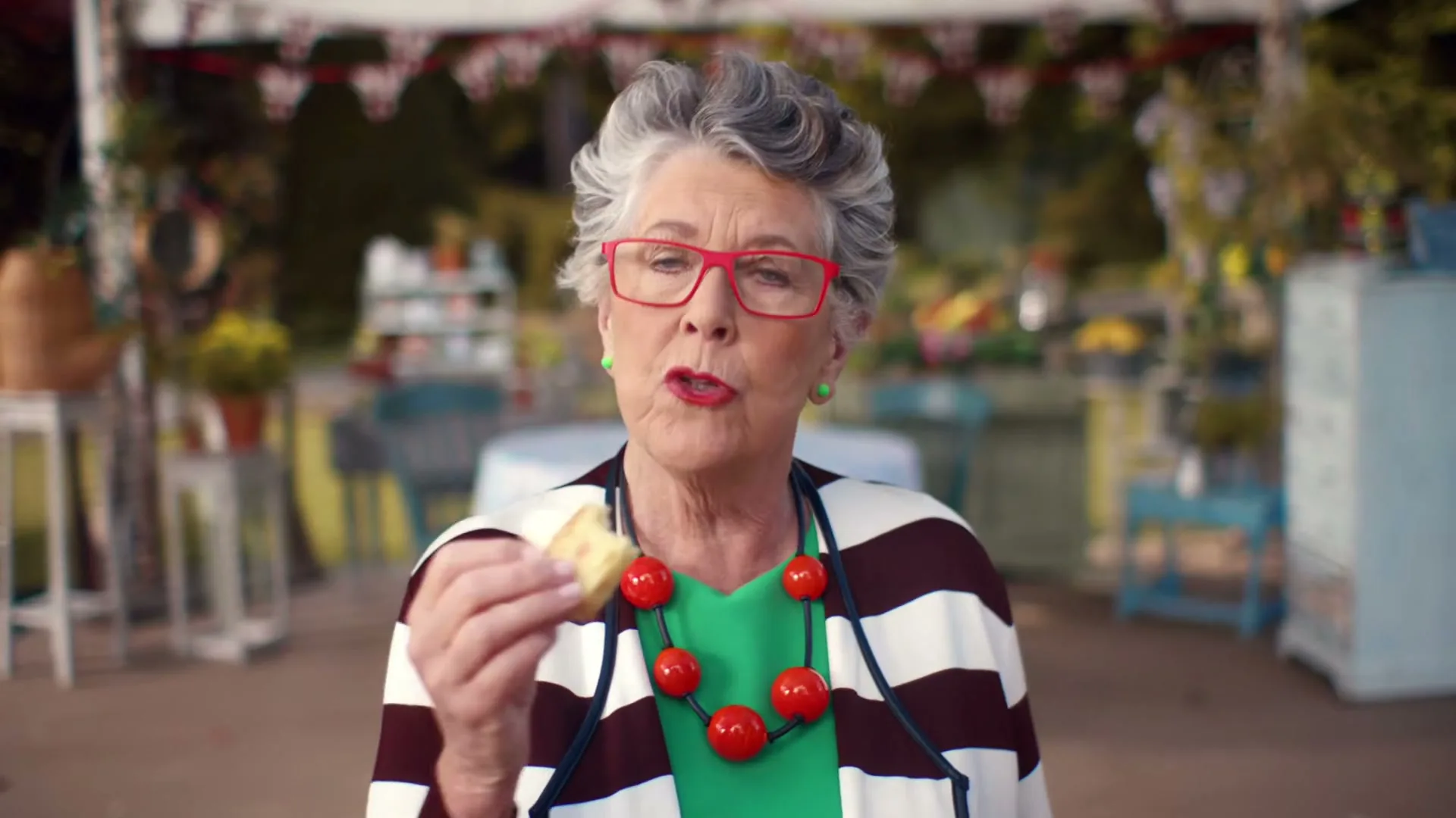 A senior woman with grey hair and red glasses eats a small cake, looking directly at the camera with a disapproving expression in an outdoor setting.