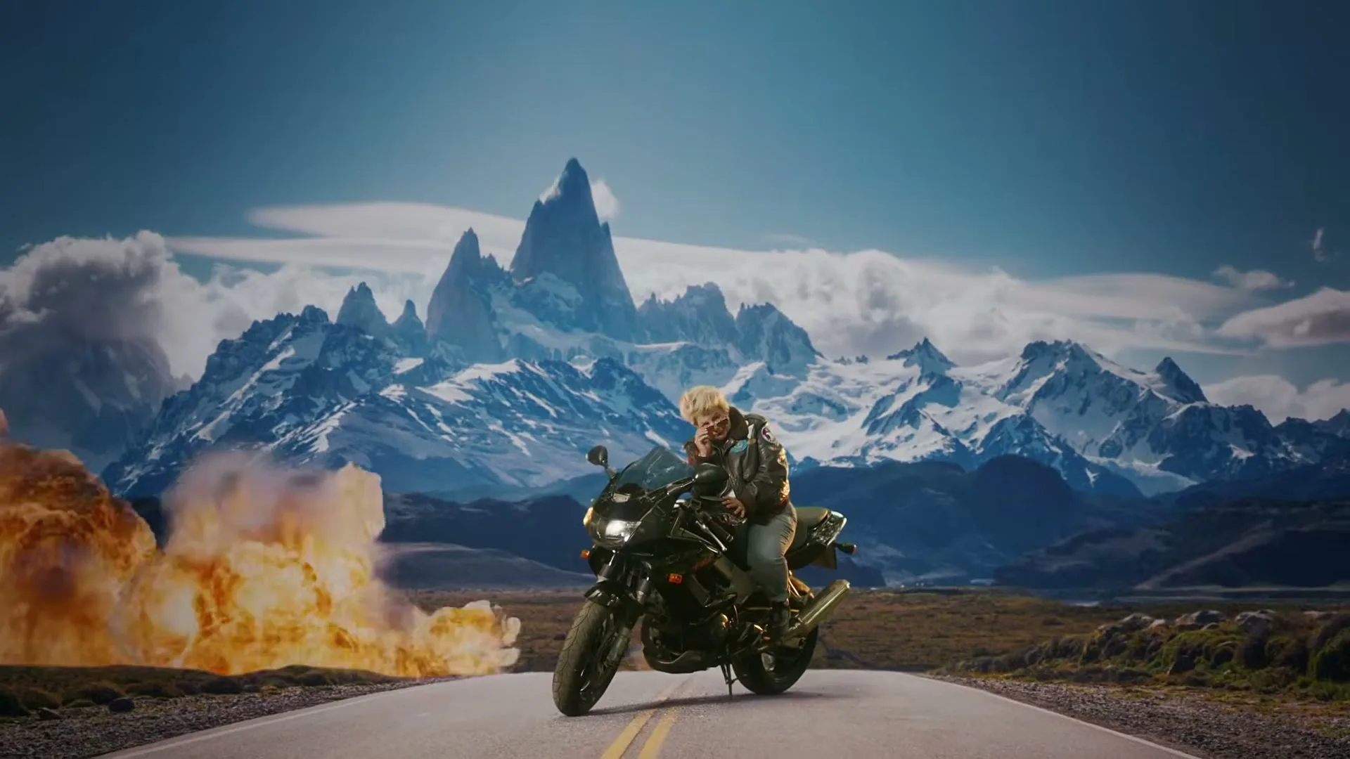 A woman with blonde hair and a leather jacket sits on a motorcycle on a road, with a massive explosion behind her and snow-capped mountains in the background.