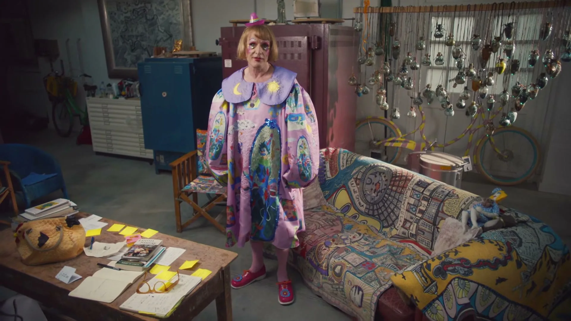 A person in a flamboyant, oversized purple dress with moon and sun motifs stands in a cluttered art studio, looking directly at the camera.