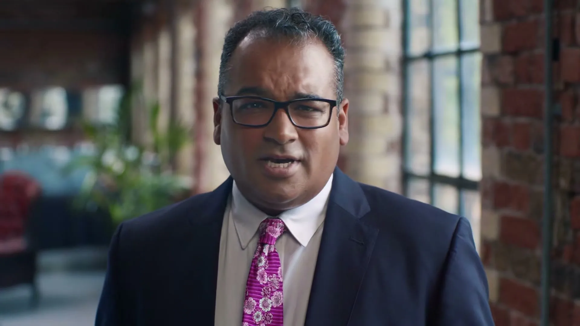 A man in his 40s, wearing a suit and a pink tie, speaks directly to the camera in an office setting.