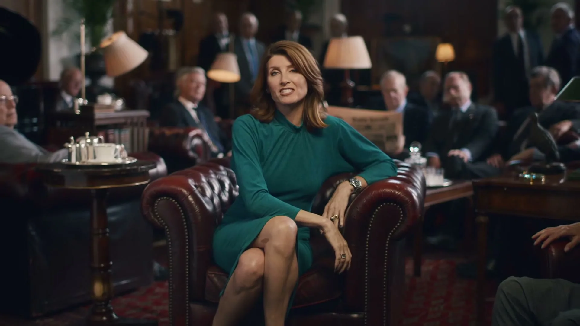 A woman in a green dress sits in a leather armchair, speaking to the camera, surrounded by men in a dimly lit, traditional club setting.
