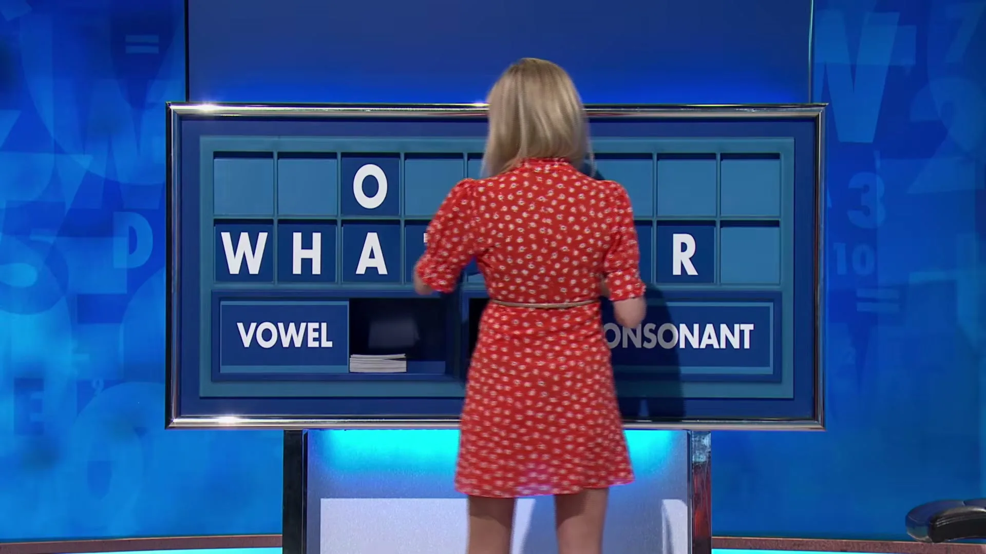 A blonde woman in a red dress stands with her back to the camera, selecting letters on a large blue game show board.