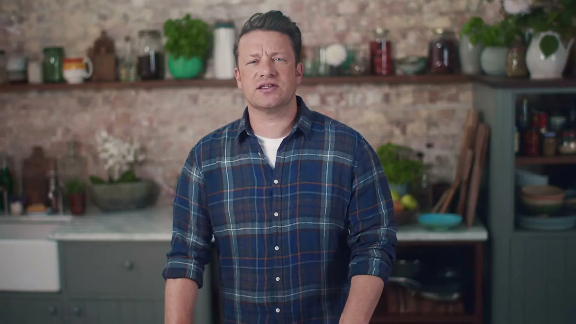 Jamie Oliver, a male in his 40s, wearing a blue plaid shirt, stands in a rustic kitchen, looking annoyed as he reacts to a comment.