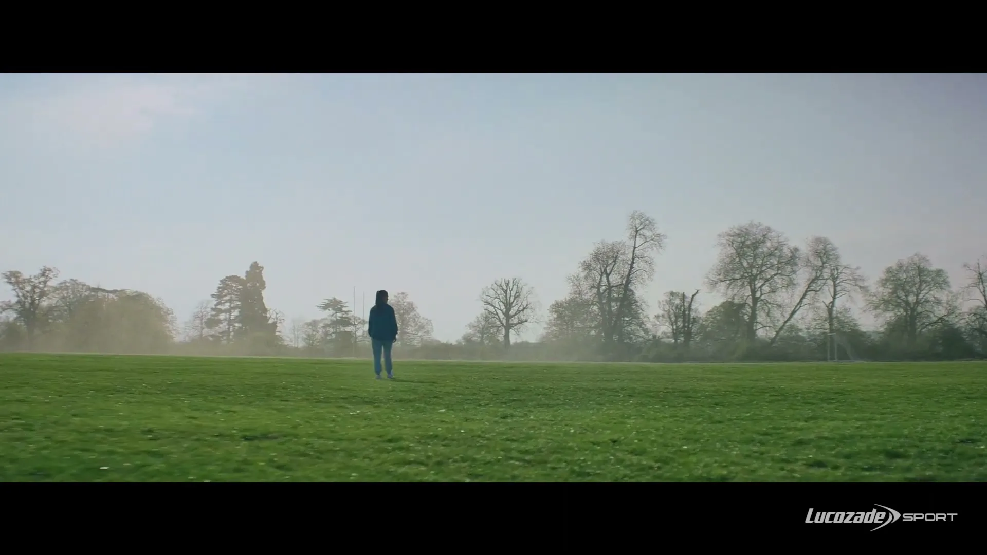 A young female soccer player walks across a wide-open green field with trees in the background under a bright sky.