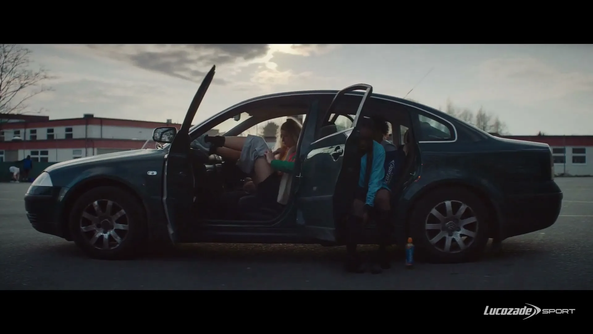 Three young women in athletic wear are getting out of a dark sedan in a school parking lot at dusk, with a building in the background.