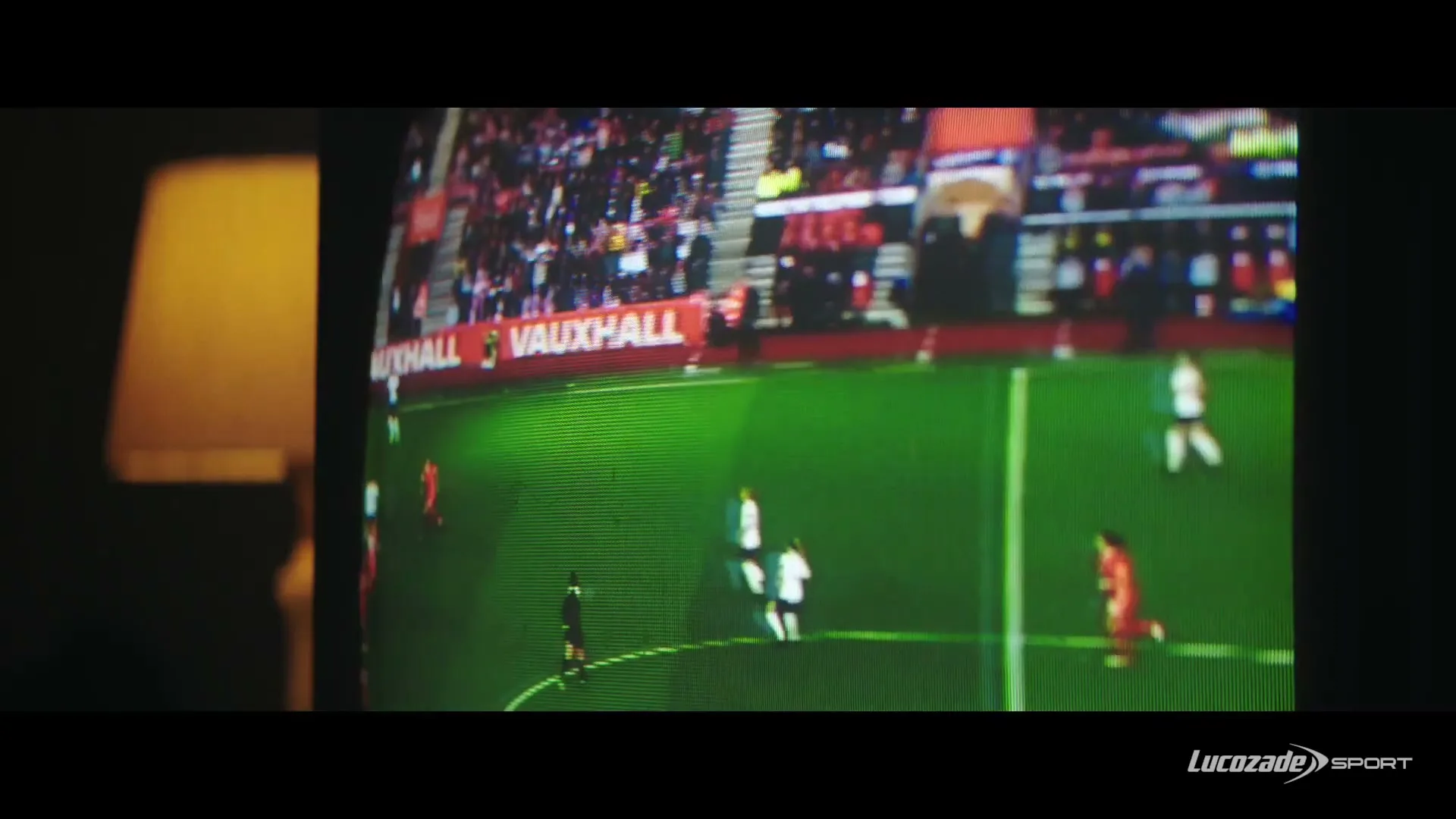 A wide shot of a soccer game on a TV screen, with players in red and white jerseys on a green field, and spectators in the background.