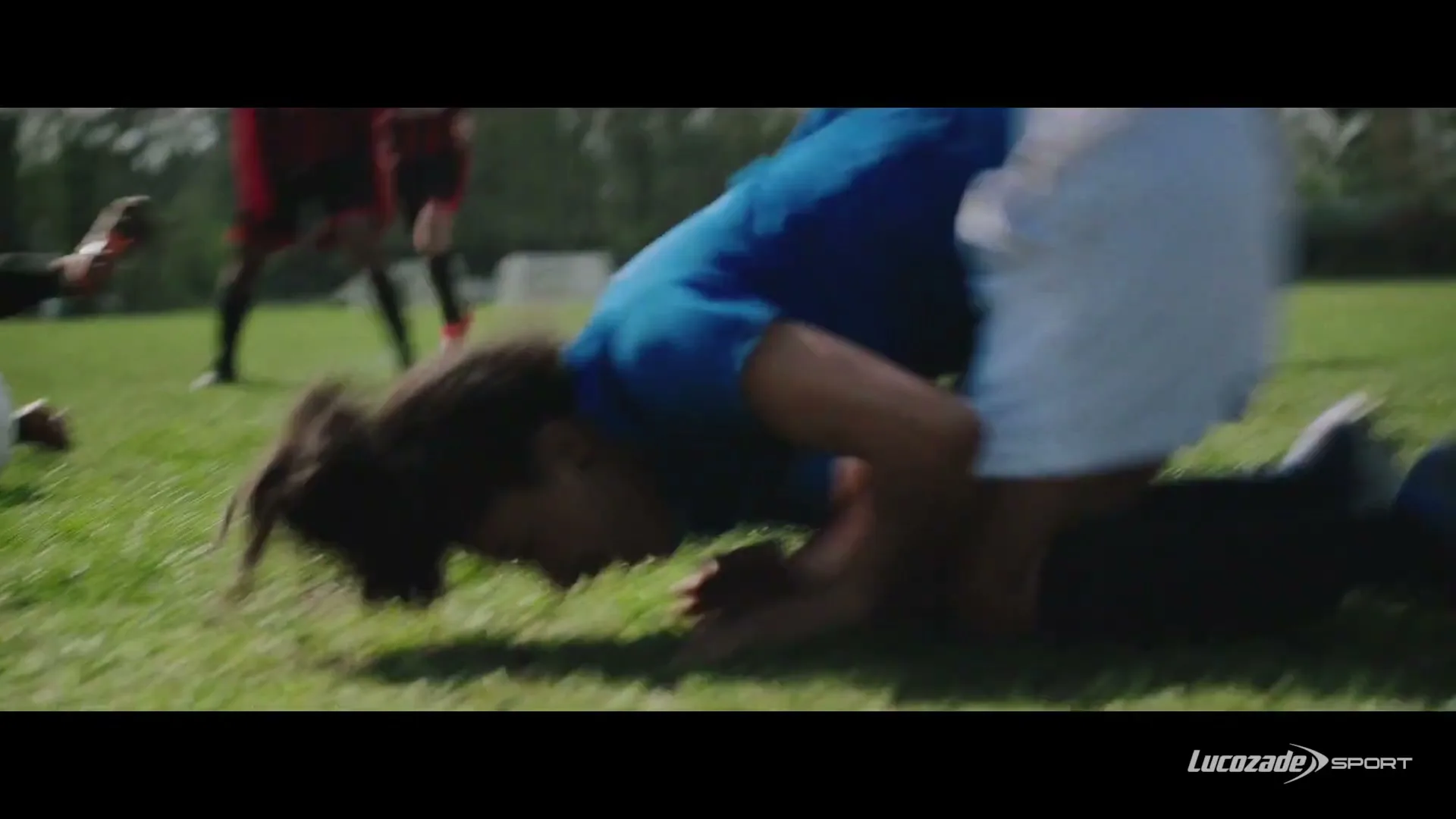 A female soccer player in a blue jersey is tackled by an opponent in a red and black jersey on a grassy field during a game.