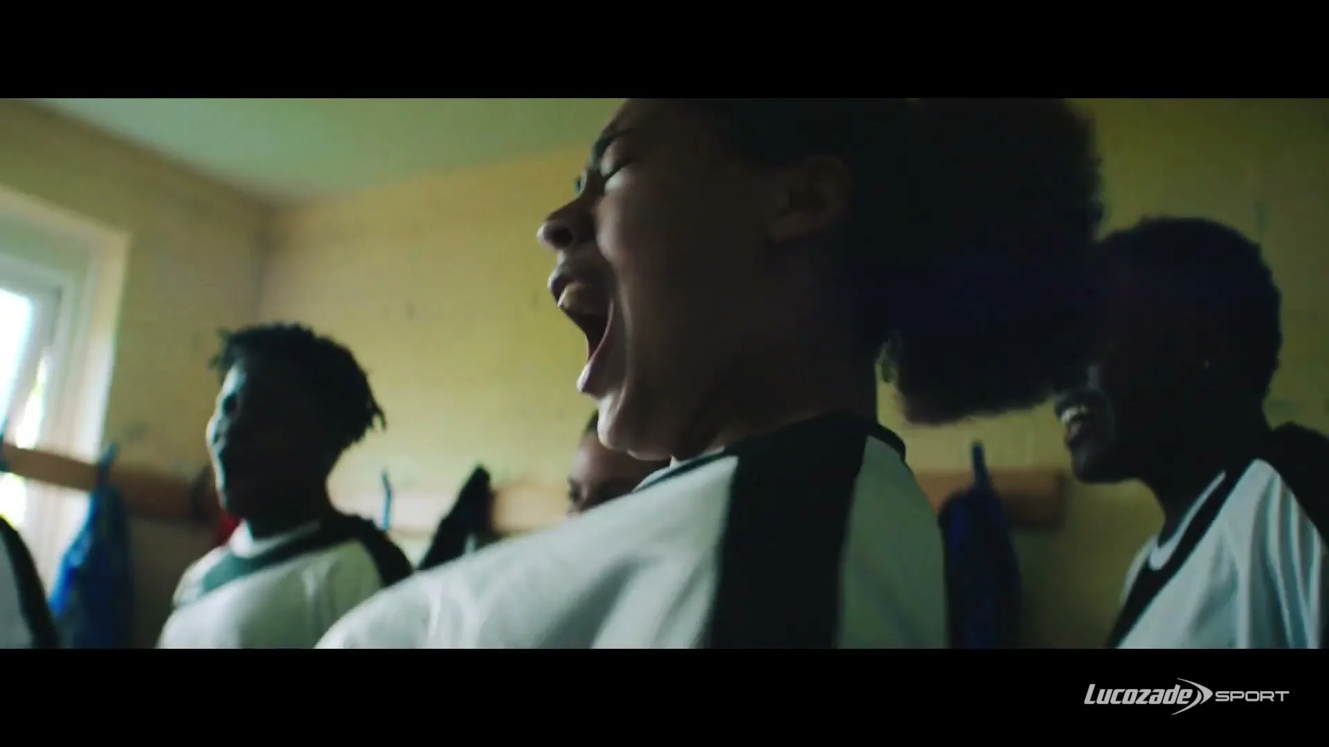 A young female soccer player with curly hair shouts in a locker room, surrounded by teammates, in a medium closeup shot.