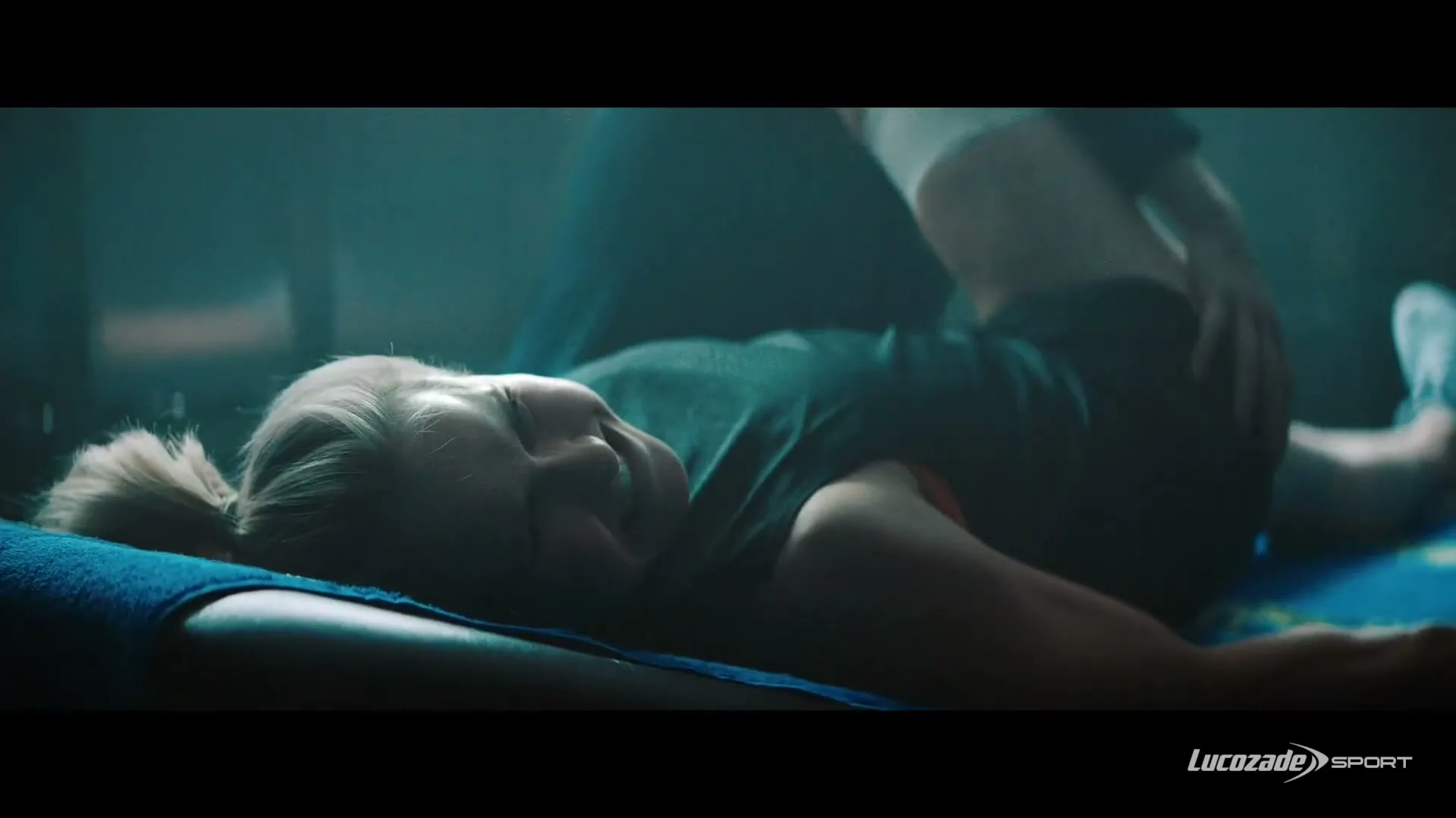 A female athlete with blonde hair in a ponytail lies on her back on a blue mat, smiling as someone stretches her leg in a dimly lit gym.