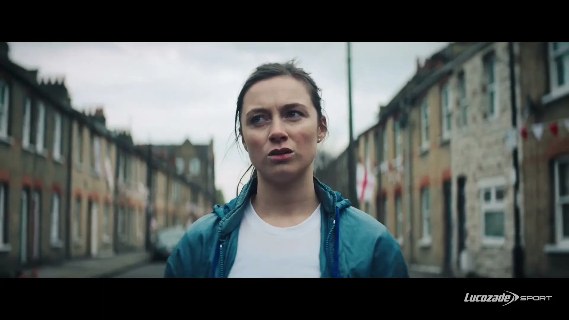 A young woman in a blue jacket and white t-shirt stands in the middle of a residential street, looking around with a concerned expression.