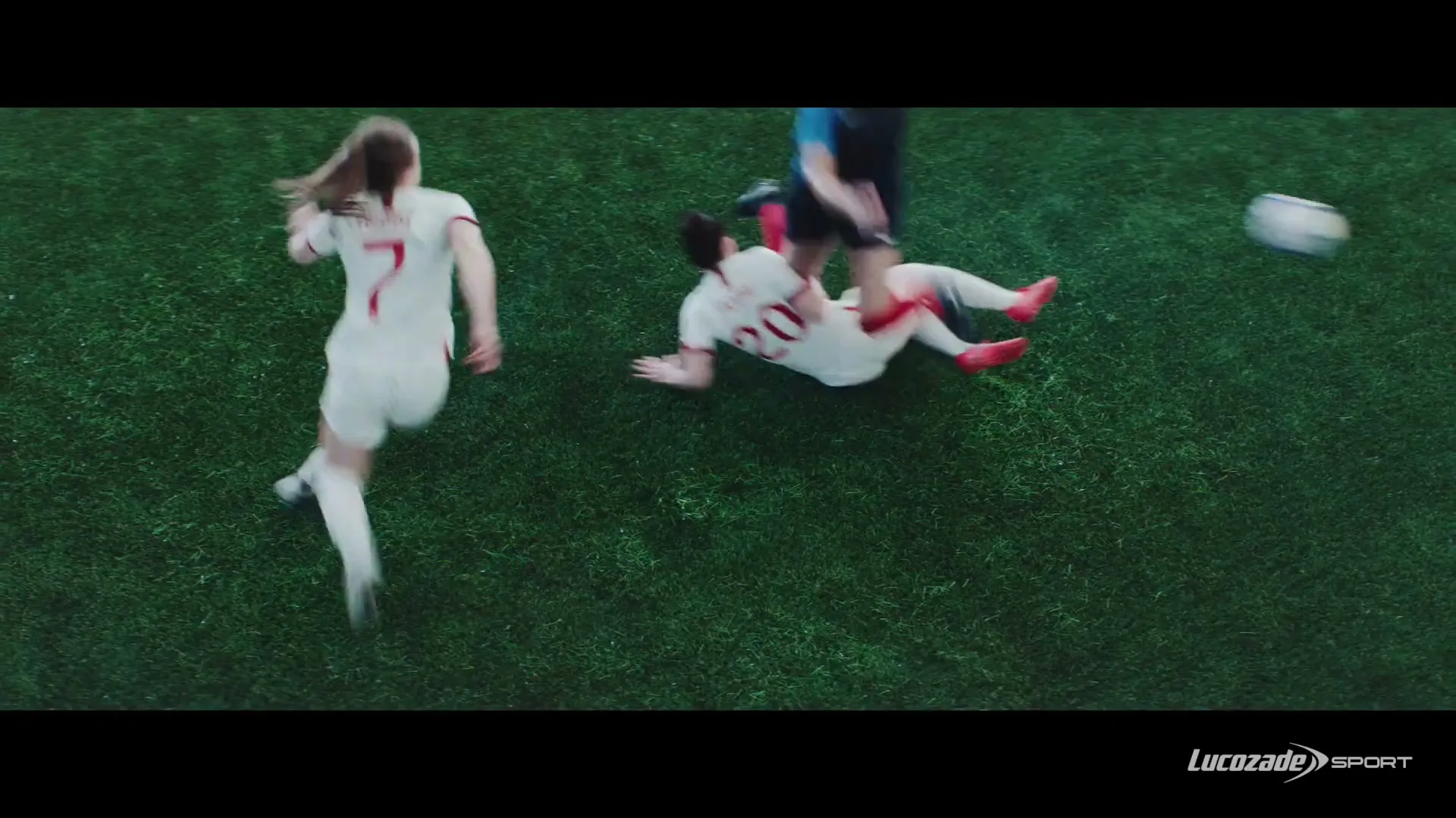 Three female soccer players in white and blue jerseys are on a green grass field, one falling, one running, and one tackling the ball.