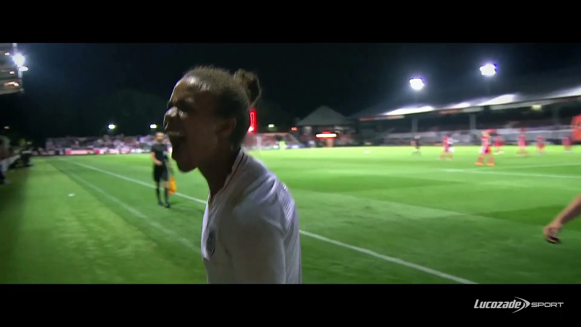 A female soccer player in a white jersey with her hair in a bun screams in excitement on a floodlit soccer field at night.