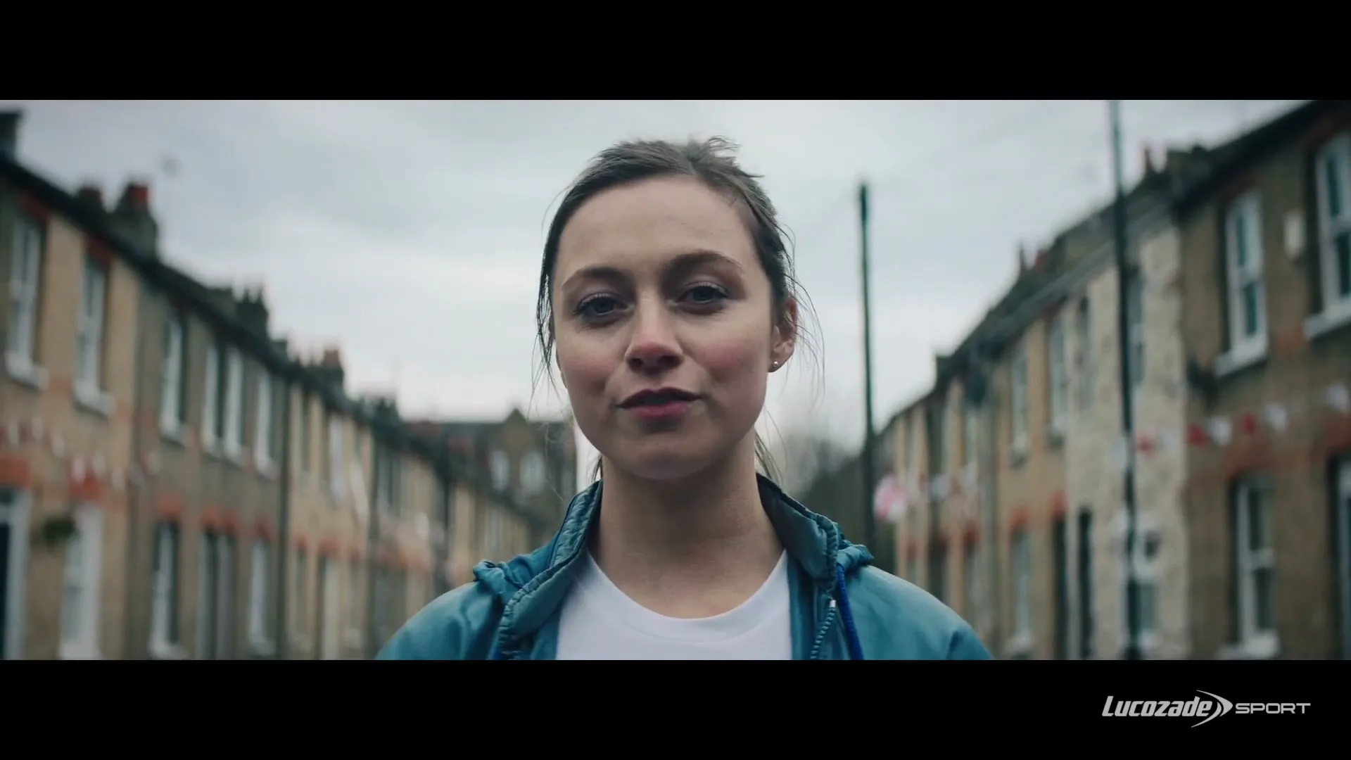 A young woman in a blue jacket and white t-shirt stands in the middle of a residential street, looking directly at the camera with a slight smile.