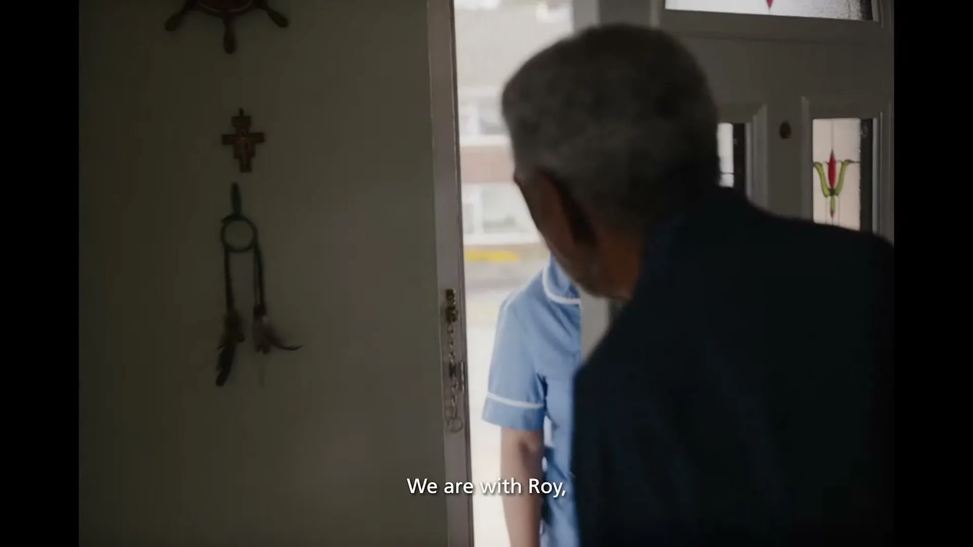An older man in a blue shirt opens his front door to reveal a smiling female nurse in a blue uniform, standing outside his home.