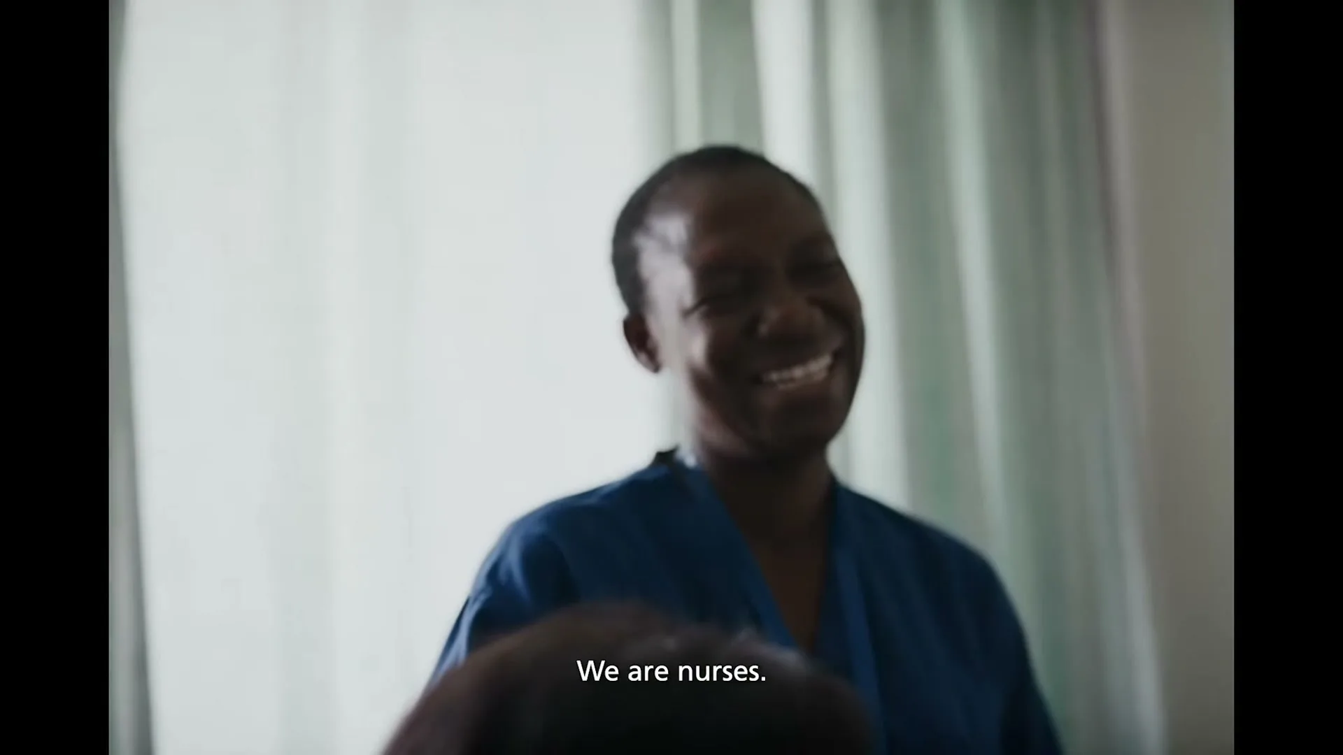 A smiling Black female nurse in blue scrubs looks at someone off-camera in a hospital room.
