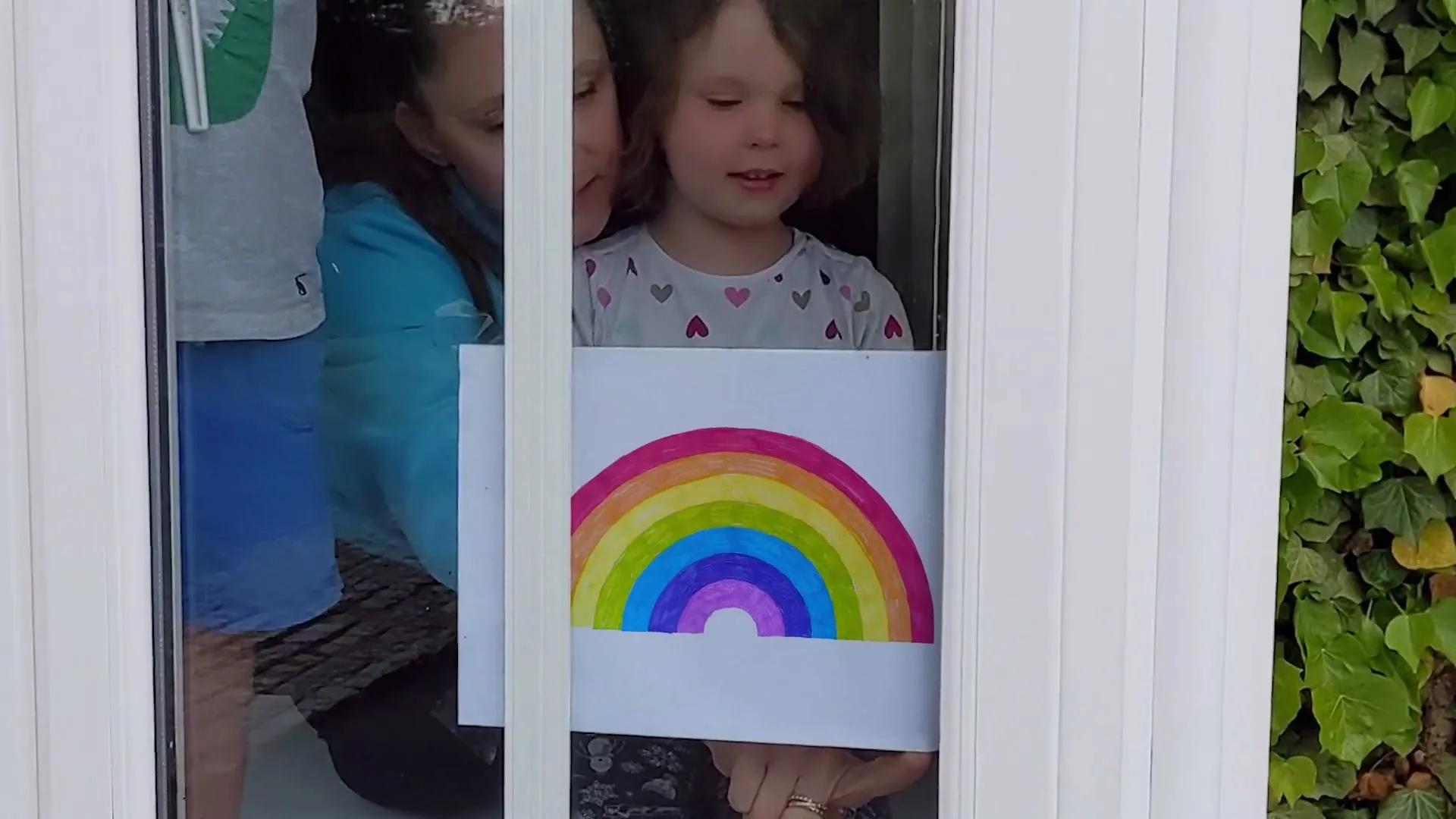A young girl with curly hair holds up a rainbow drawing in a window, with another child and adult partially visible behind her.